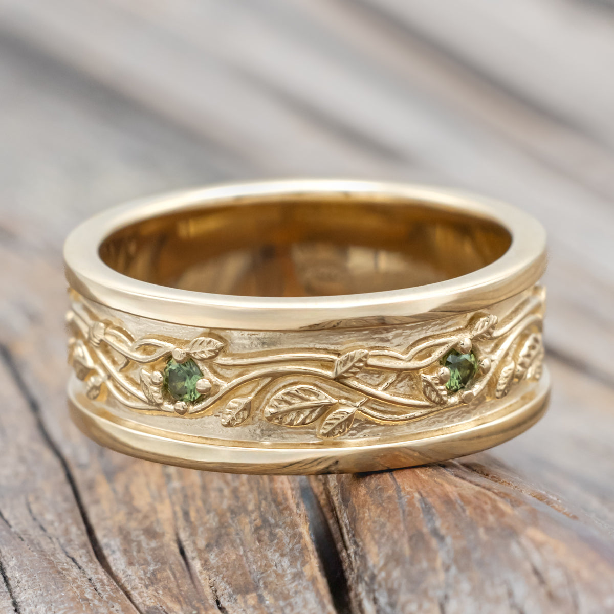 Yellow gold band with vine filigree and green sapphire accents against a blurred wood background