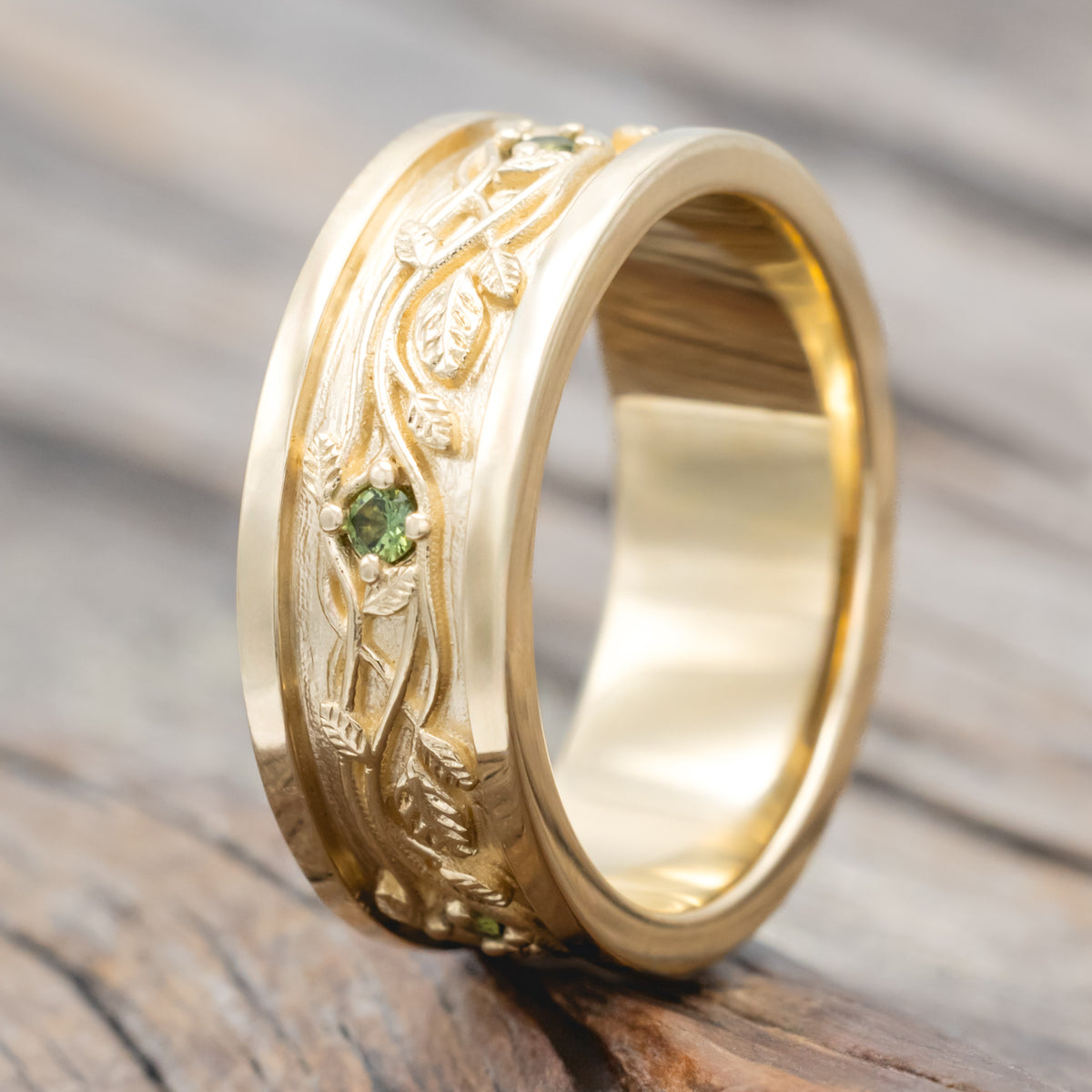 Yellow gold band with vine filigree and green sapphire accents against a blurred wood background