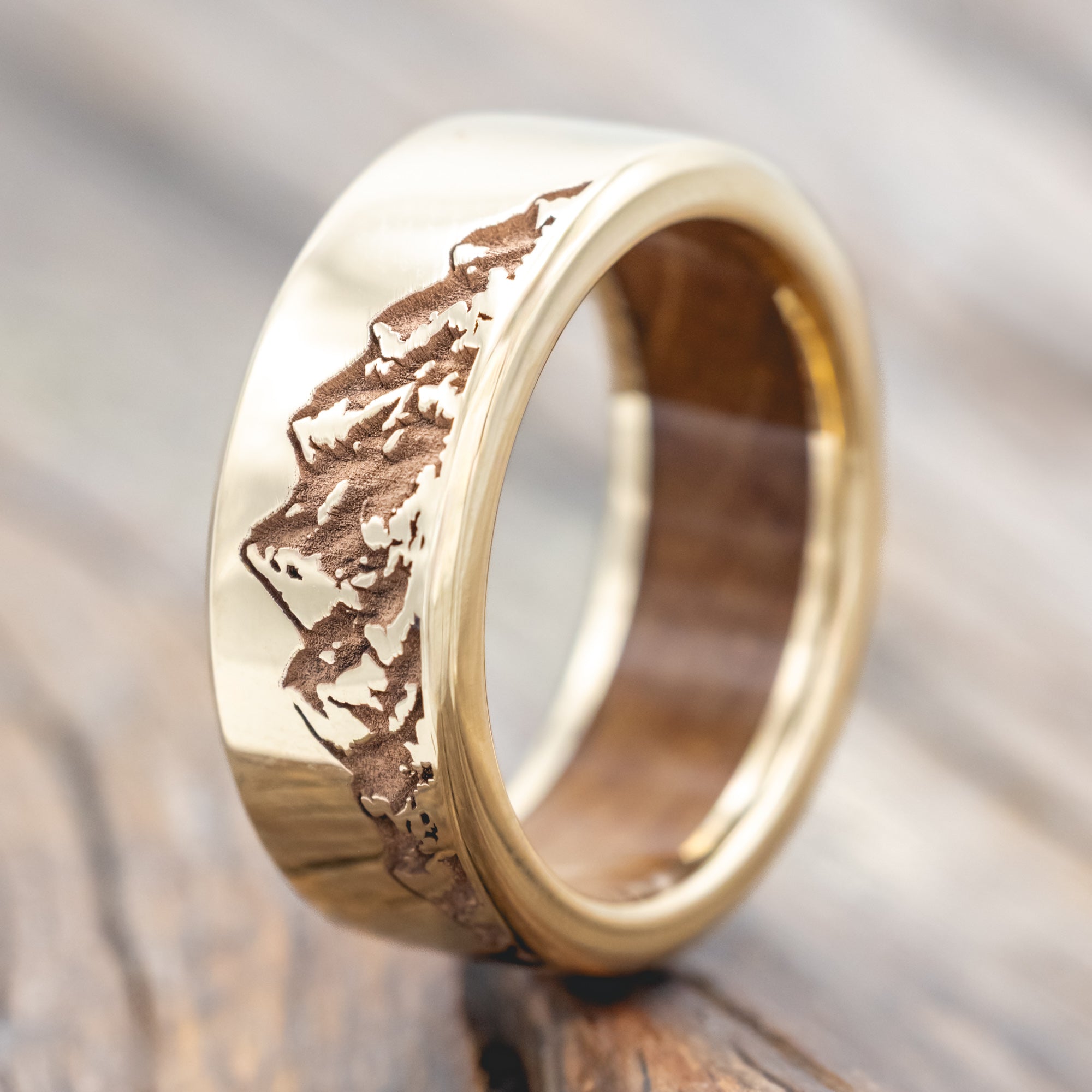 Gold ring with mountain engraving and whiskey barrel lining on a wooden surface