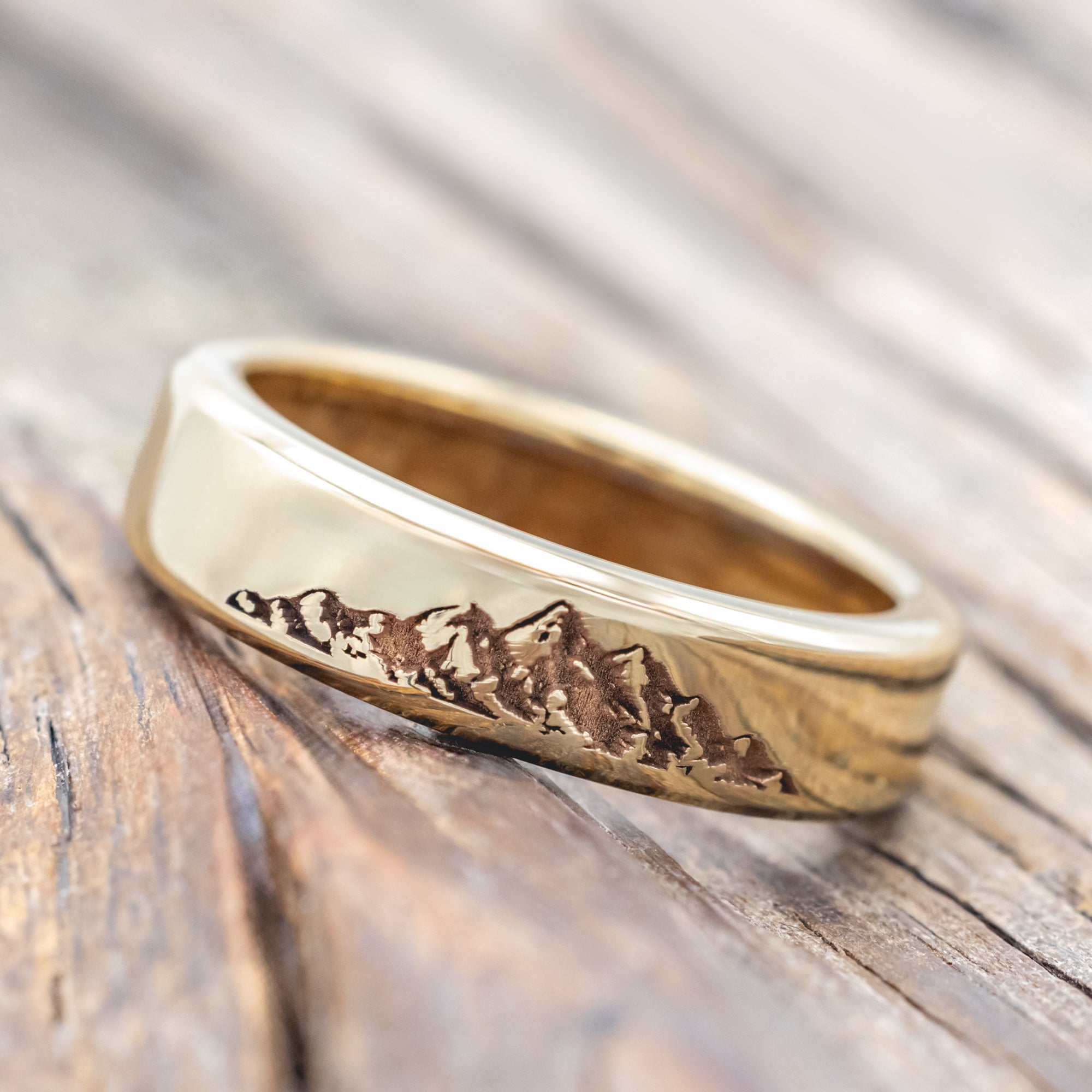 Gold ring with mountain engraving and whiskey barrel lining on a wooden surface