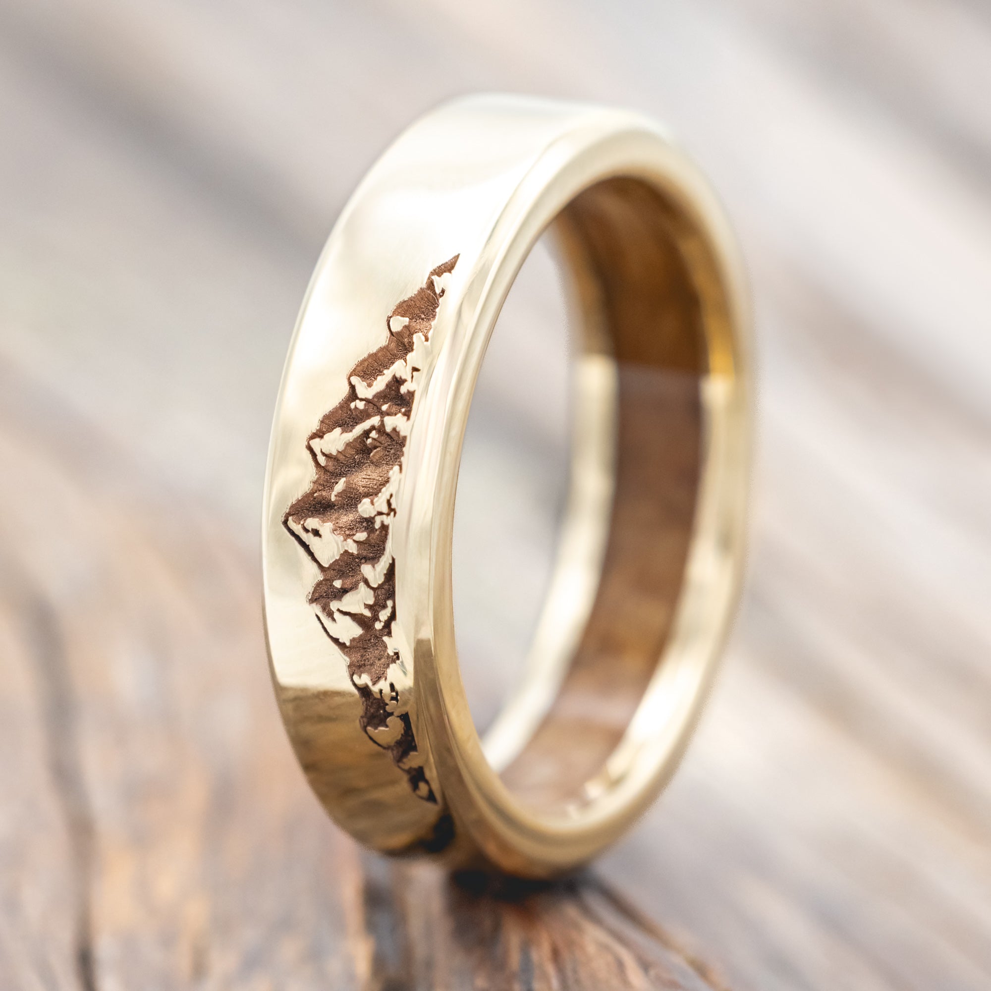 Gold ring with mountain engraving and whiskey barrel lining on a wooden surface