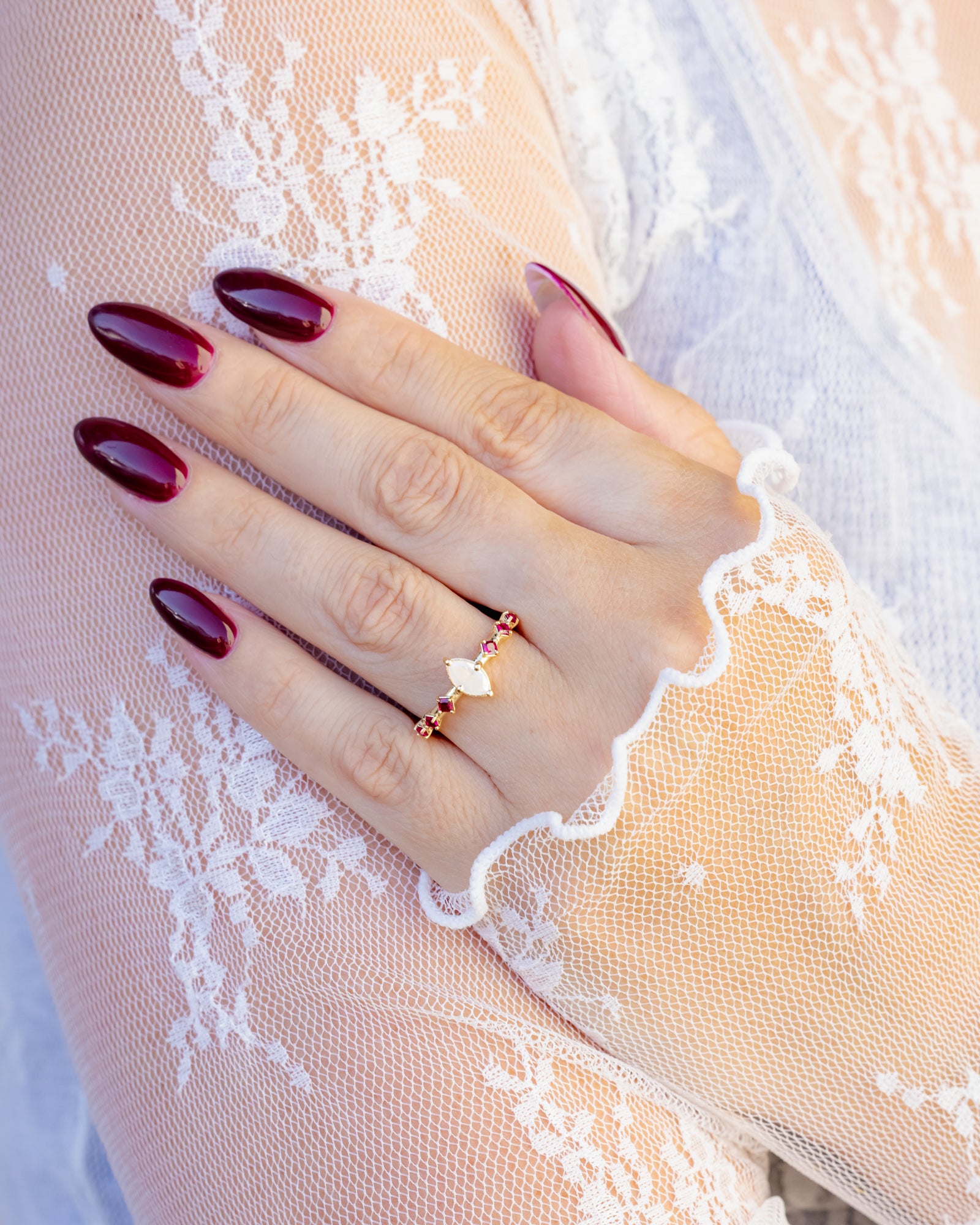 Hand with maroon nail polish wearing a delicate diamond ring with ruby accents on a lace garment