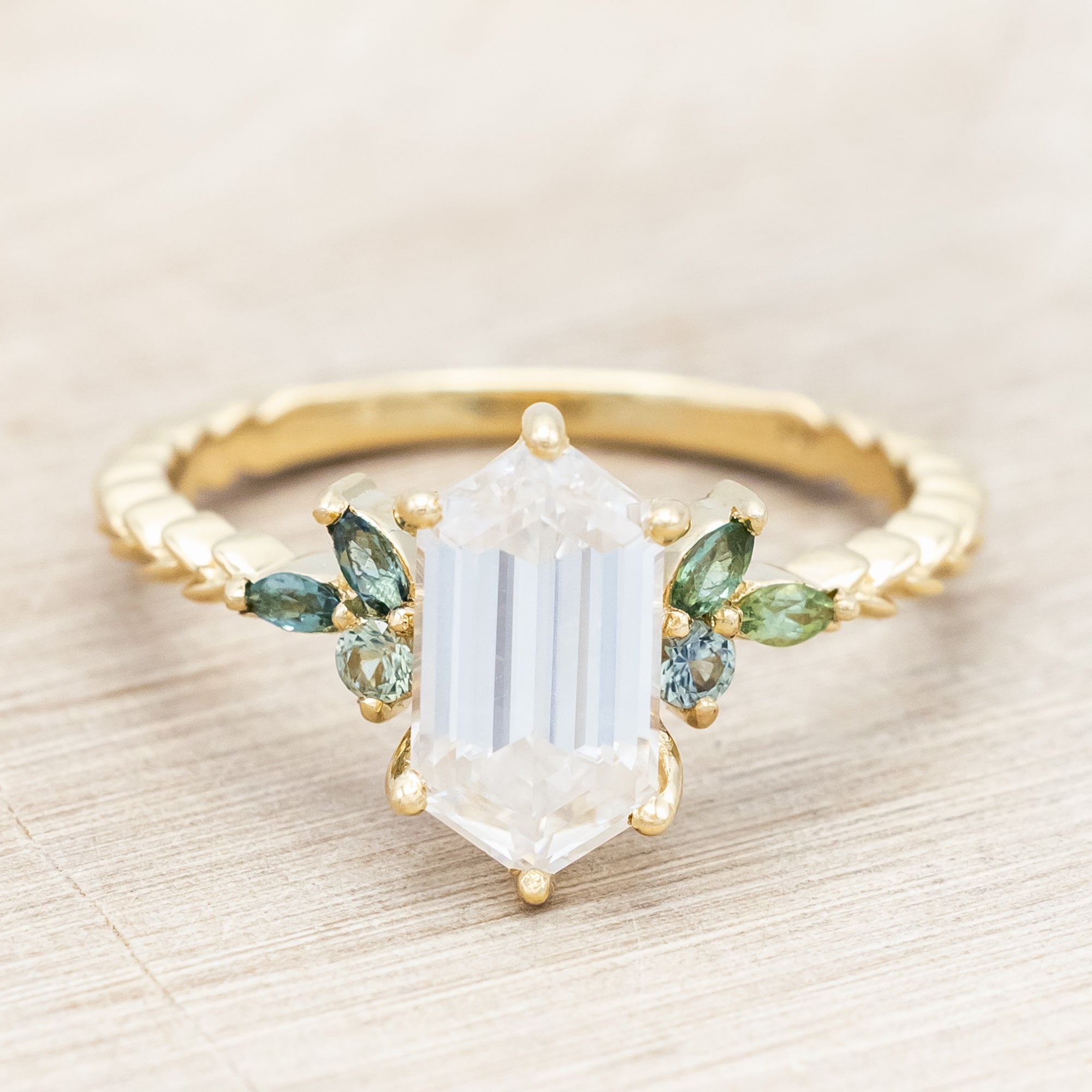 Gold ring with hexagon-cut diamond and green gemstone accents on a beige background