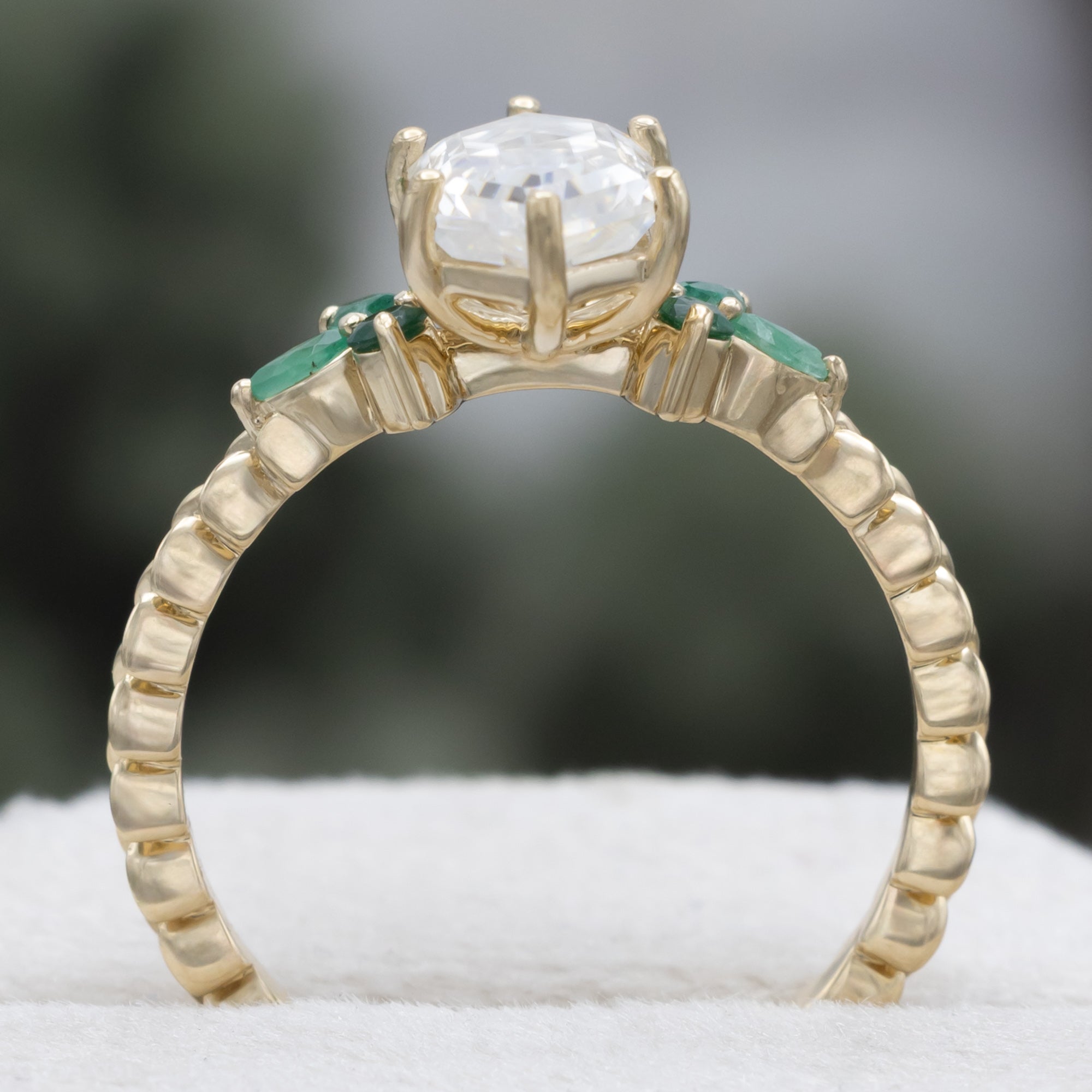 A close-up of a gold ring with a hexagon-cut moissanite and multiple green emerald accents against a blurred natural background.