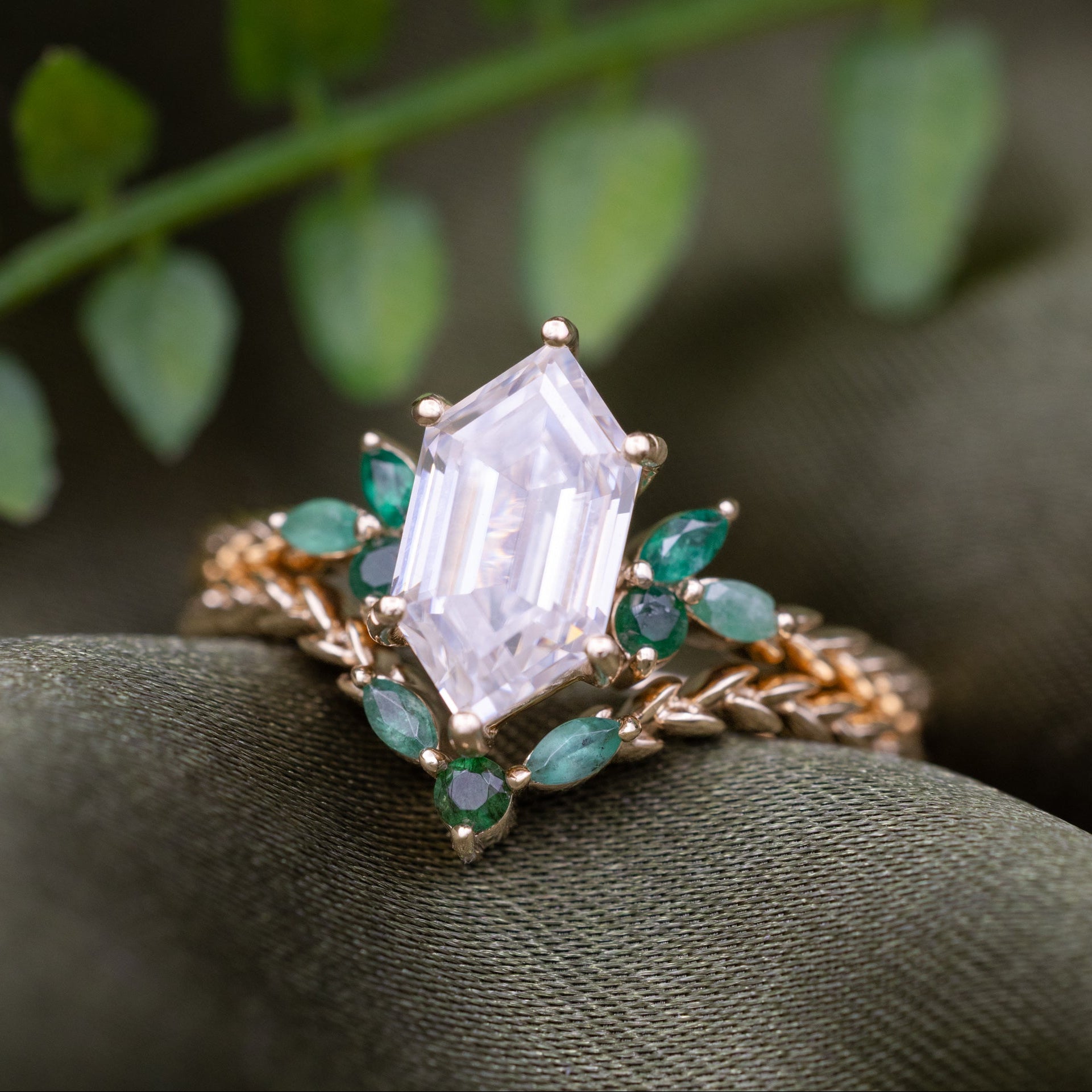 A gold ring with a hexagon Moissanite center stone and emerald accent stones, displayed on a textured olive green surface with blurred green leaves in the background.