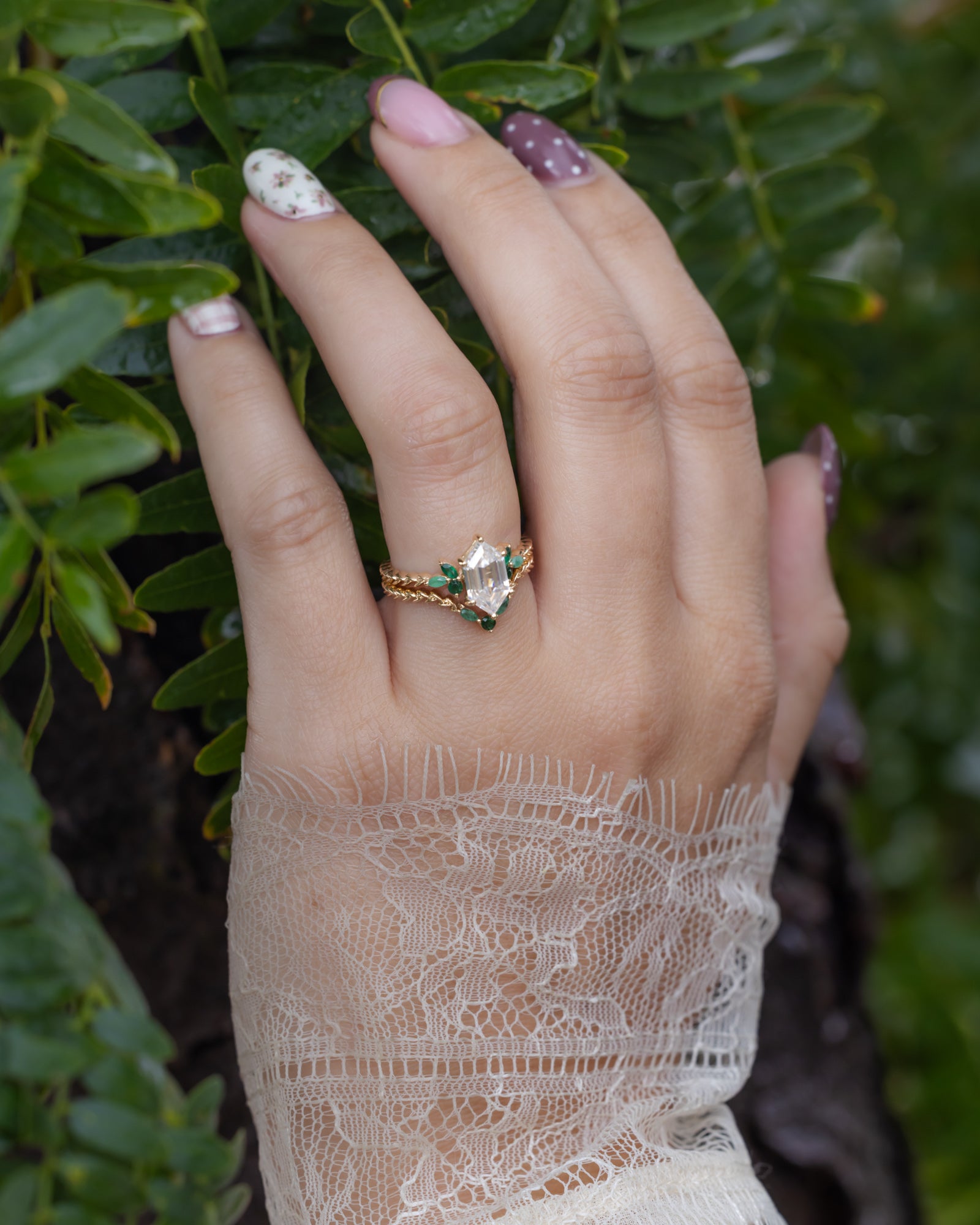 Hand with lace sleeve wearing gold ring with moissanite center stone and emerald accents against a leaf background