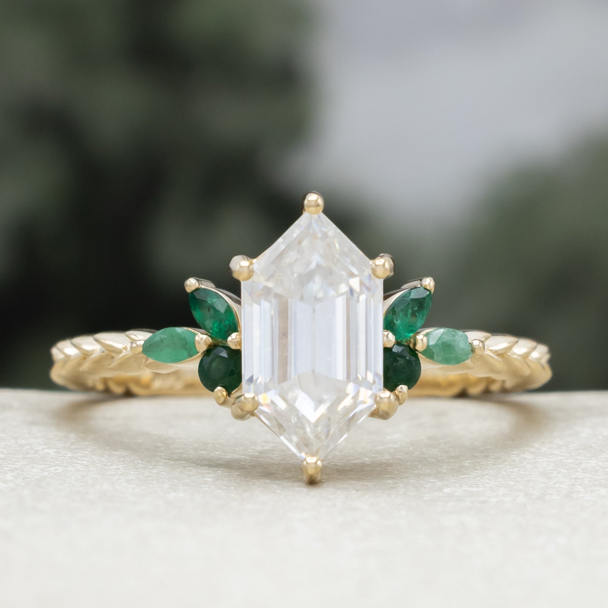 A close-up of a gold ring with a hexagon-cut moissanite and multiple green emerald accents against a blurred natural background.