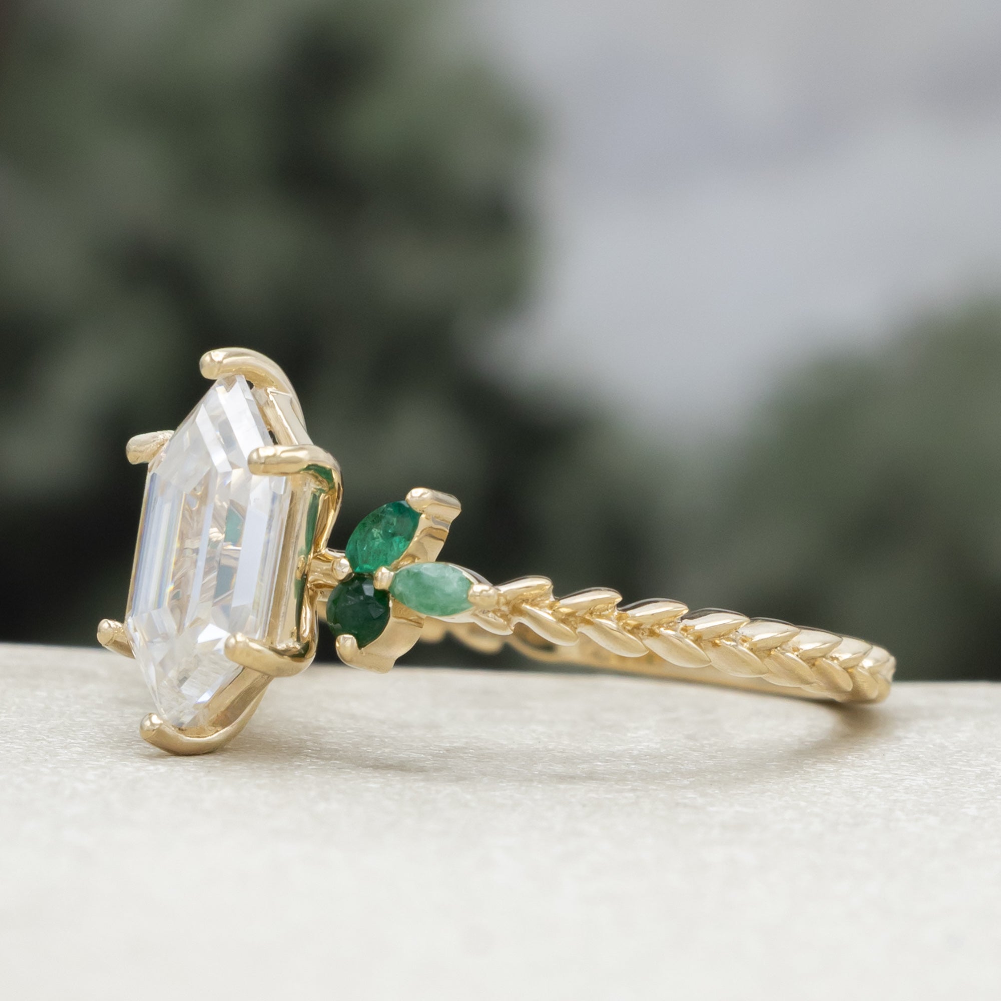 A close-up of a gold ring with a hexagon-cut moissanite and multiple green emerald accents against a blurred natural background.
