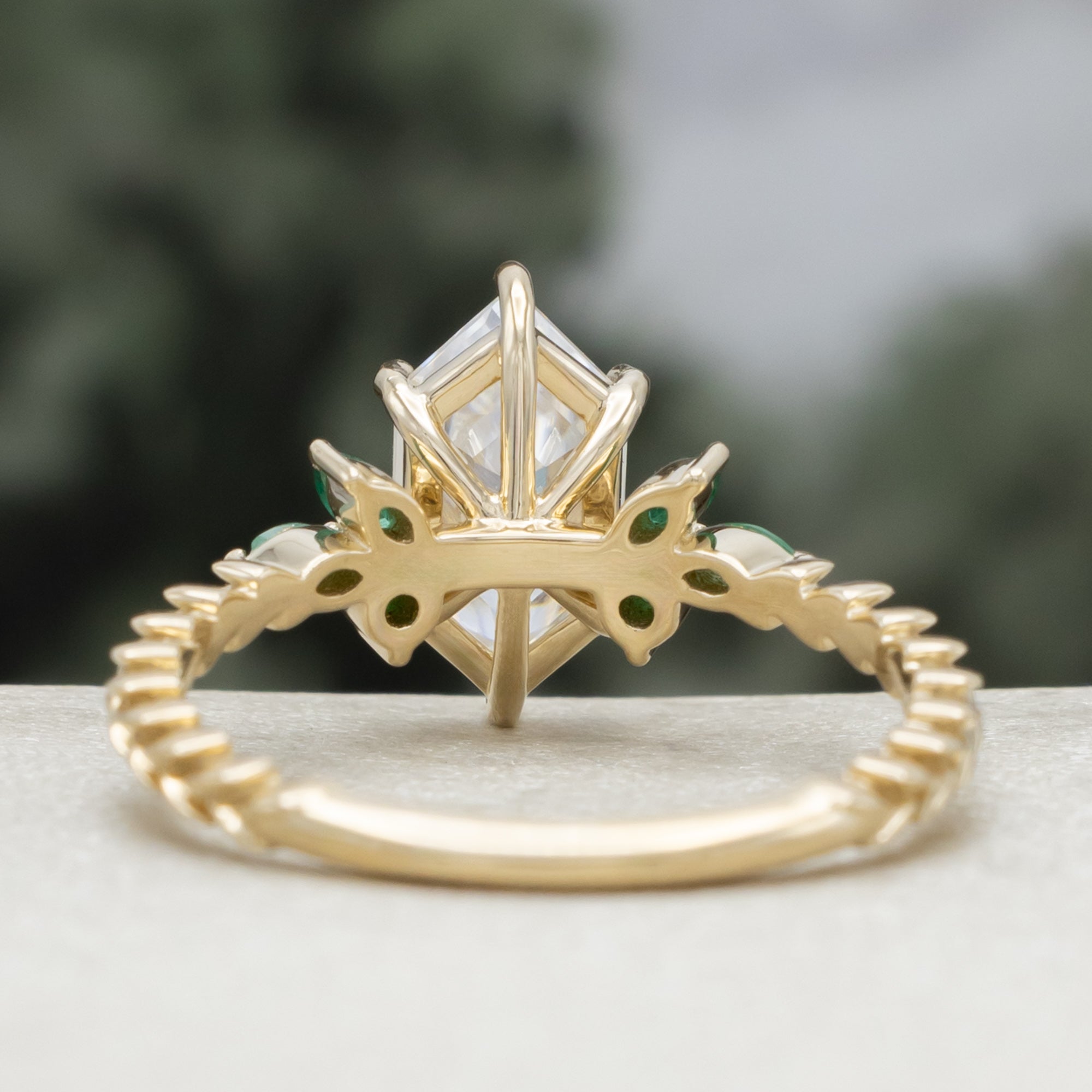 A close-up of a gold ring with a hexagon-cut moissanite and multiple green emerald accents against a blurred natural background.