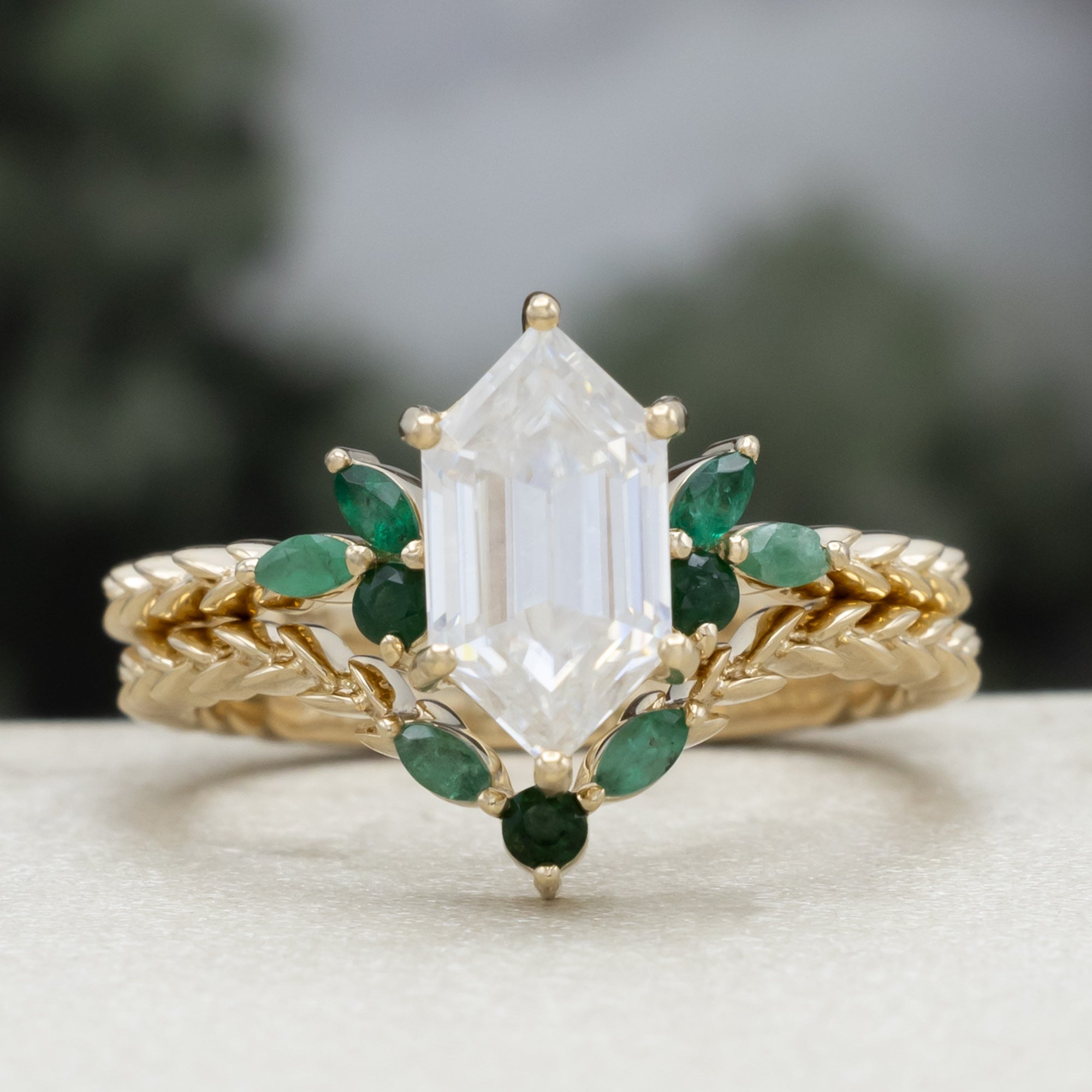 A close-up of a gold ring with a hexagon-cut moissanite and multiple green emerald accents against a blurred natural background.
