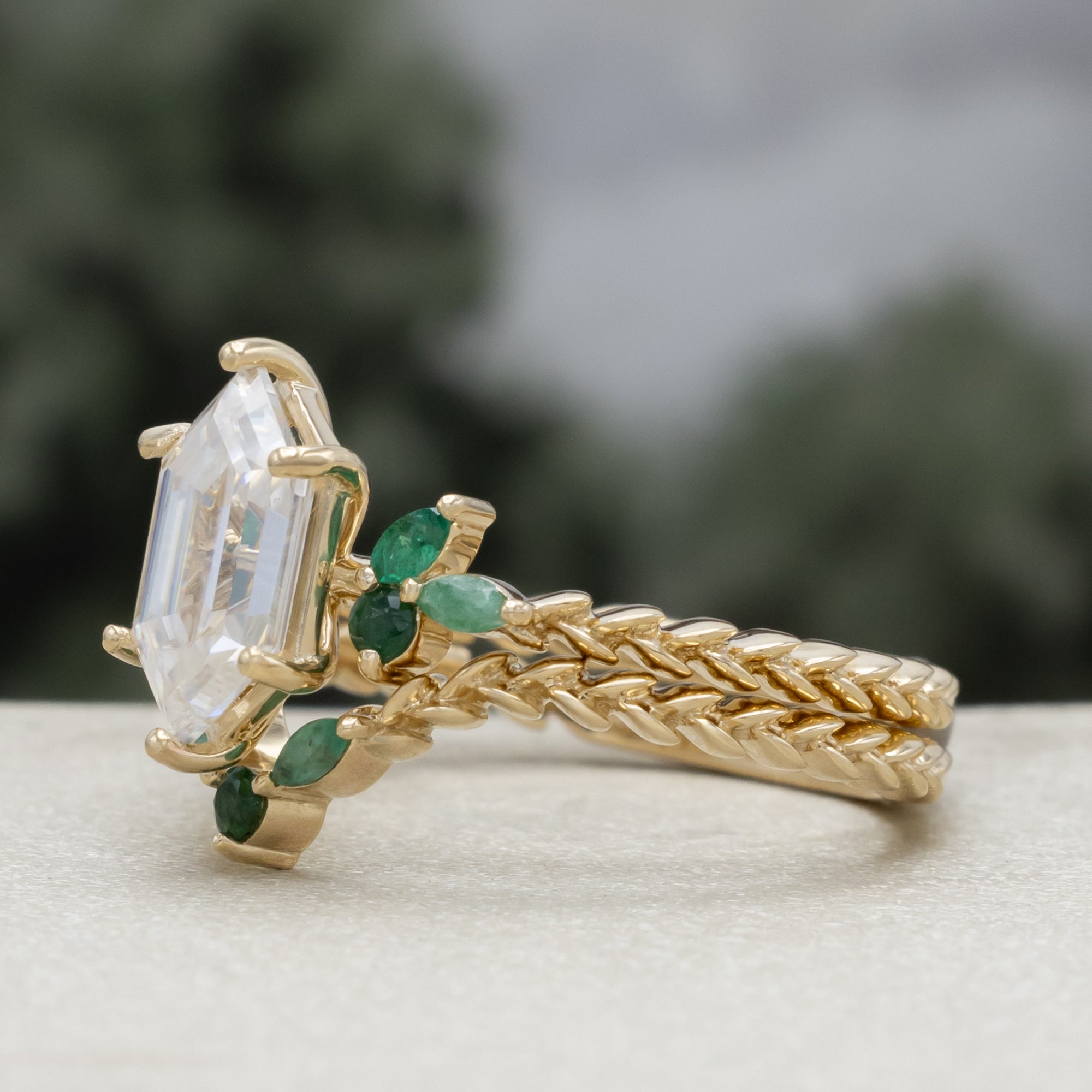 A close-up of a gold ring with a hexagon-cut moissanite and multiple green emerald accents against a blurred natural background.