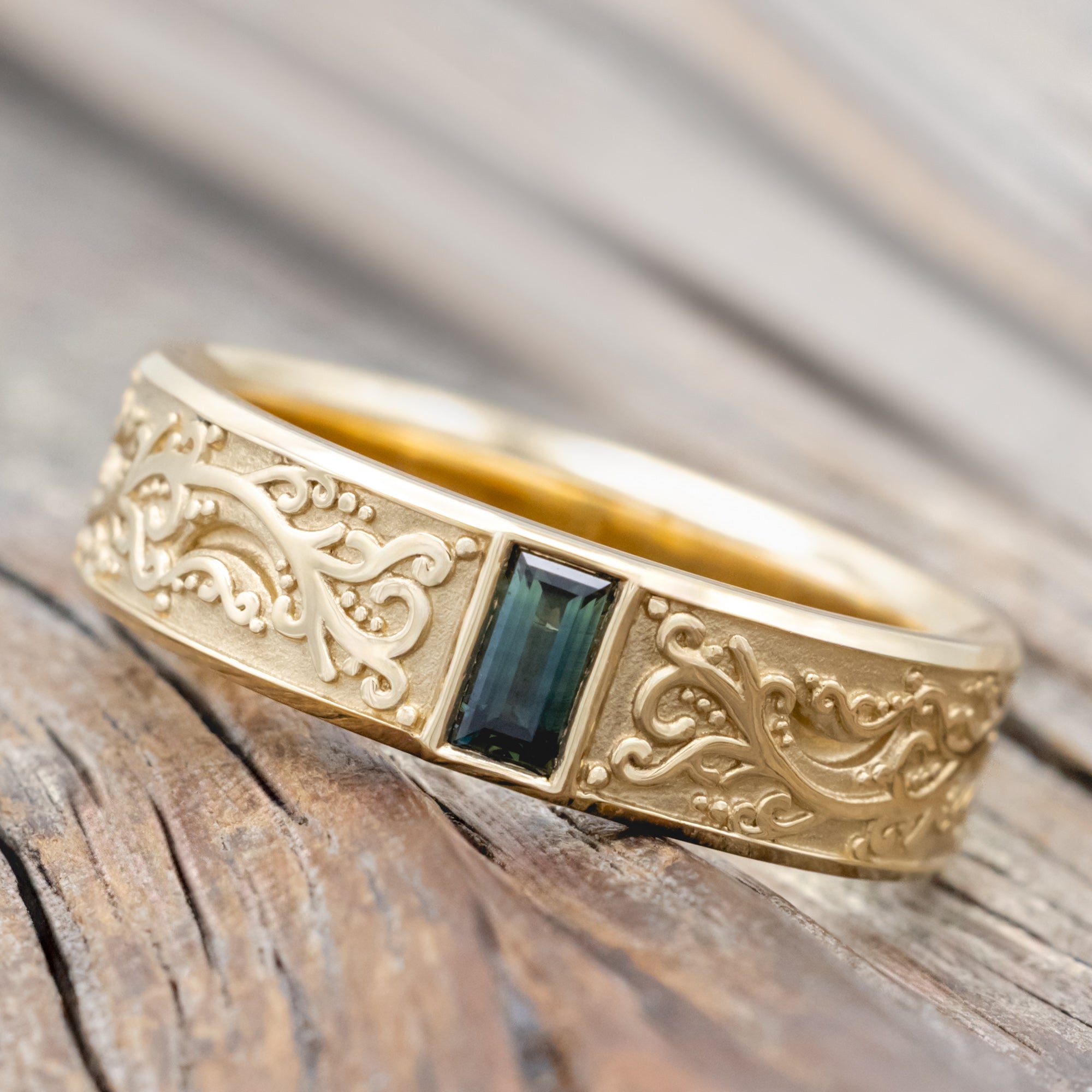 A gold ring with intricate floral engravings and a blue-green sapphire resting on a wooden surface.