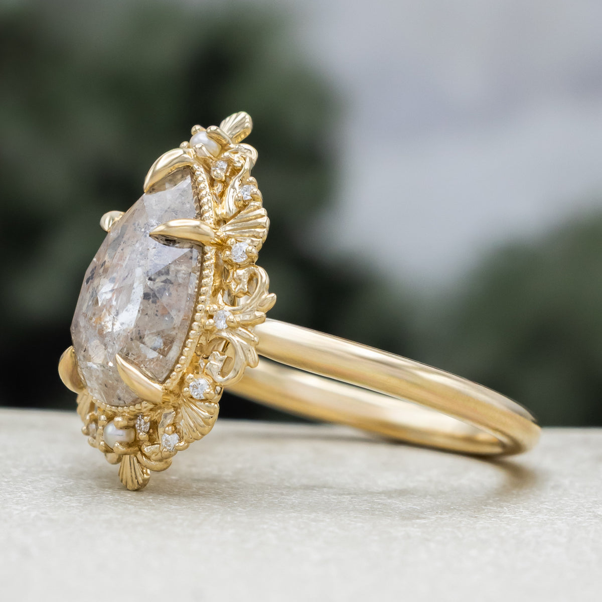 A sculptural gold ring with large pear cut salt & pepper diamond with pearl and diamond accents against a blurred green background