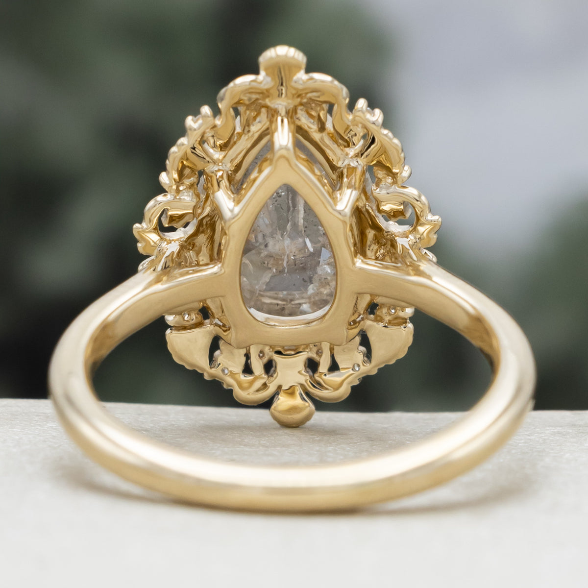 A sculptural gold ring with large pear cut salt & pepper diamond with pearl and diamond accents against a blurred green background