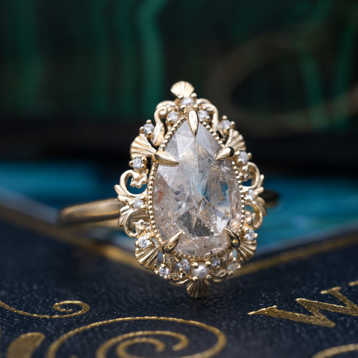A sculptural gold ring with large pear cut salt & pepper diamond with pearl and diamond accents against a dark ornate background