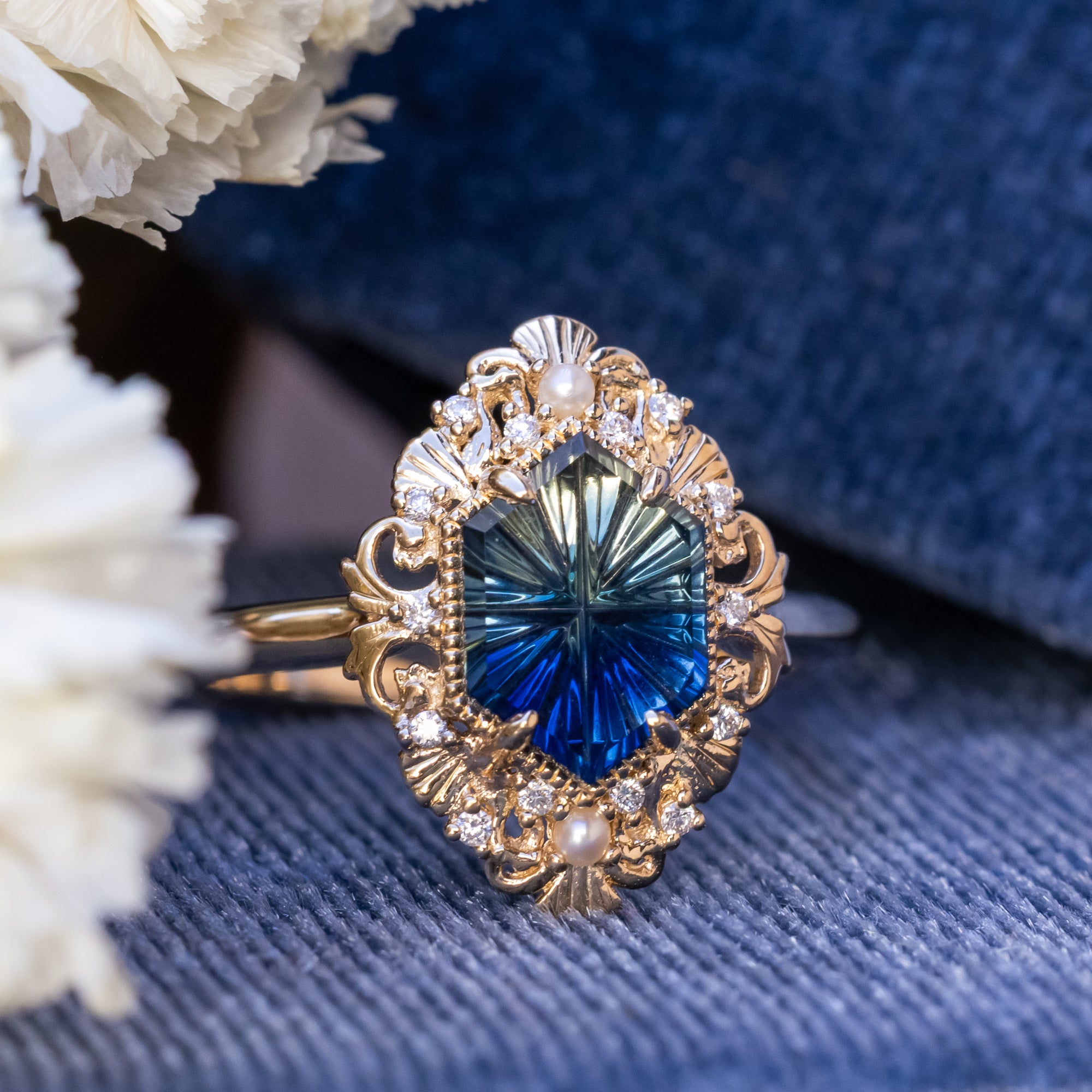 Gold ring with a blue sapphire on a textured blue surface with white flowers.