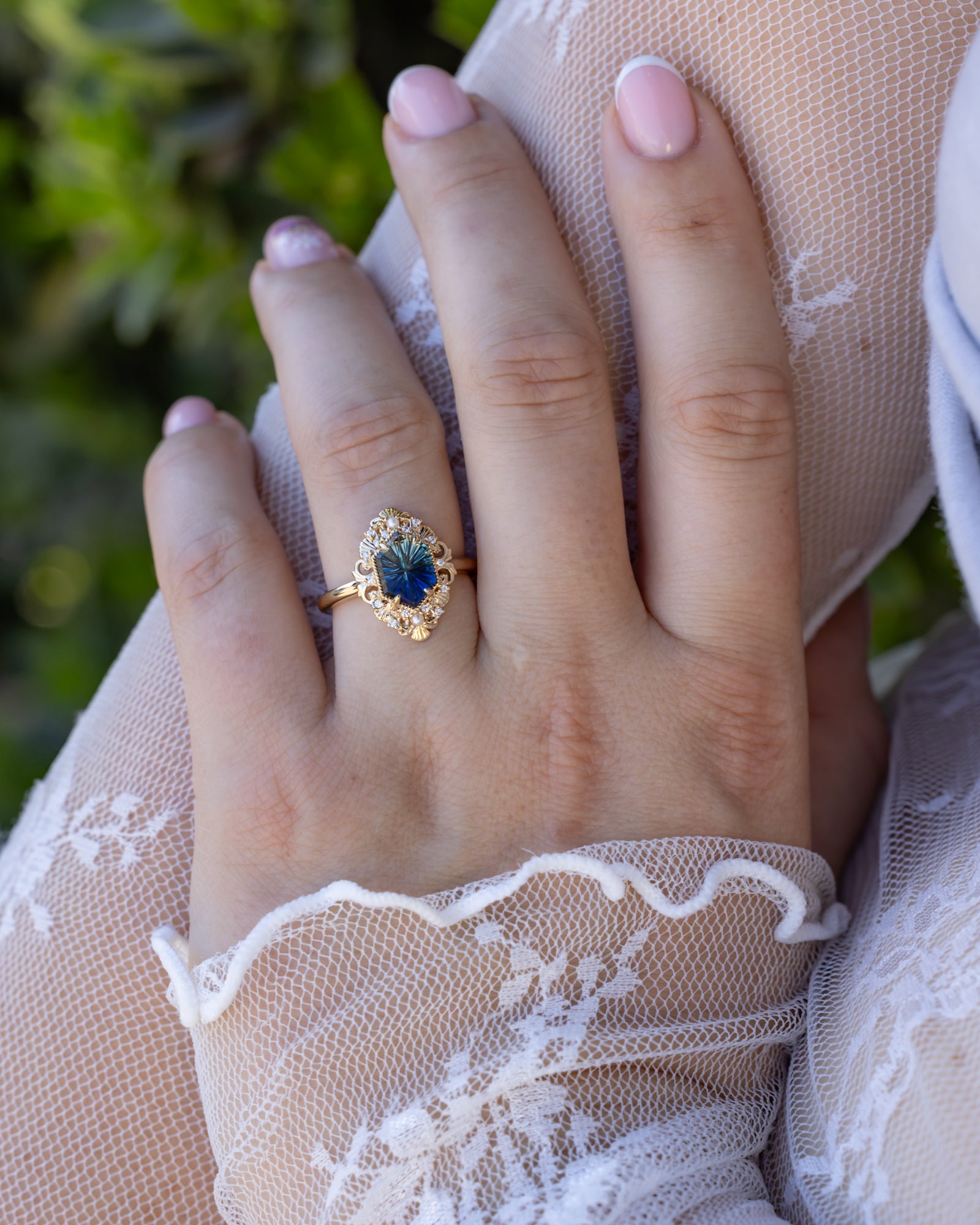 Hand wearing a gold ring with a blue sapphire against a blurred green background