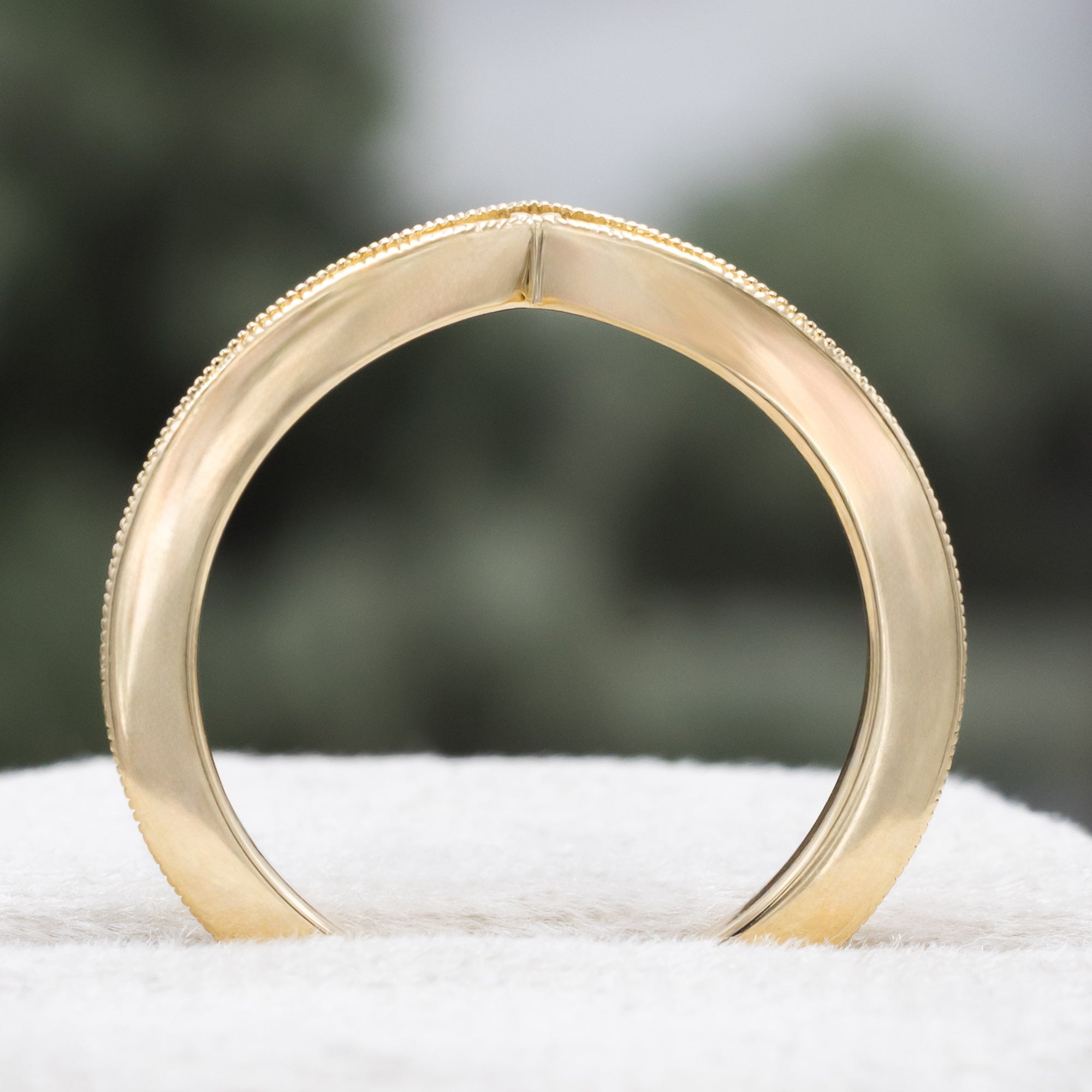 A gold V-shaped ring with leaf filigree, on a textured surface against a blurred green background
