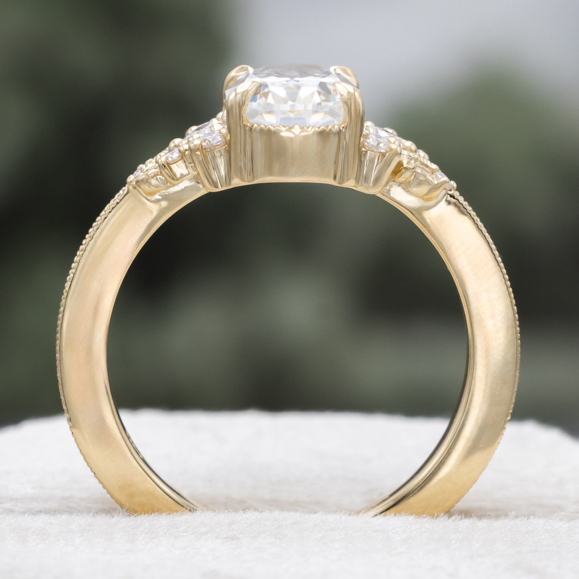 A gold engagement ring with an oval Moissanite and diamond accents, against a blurred green background