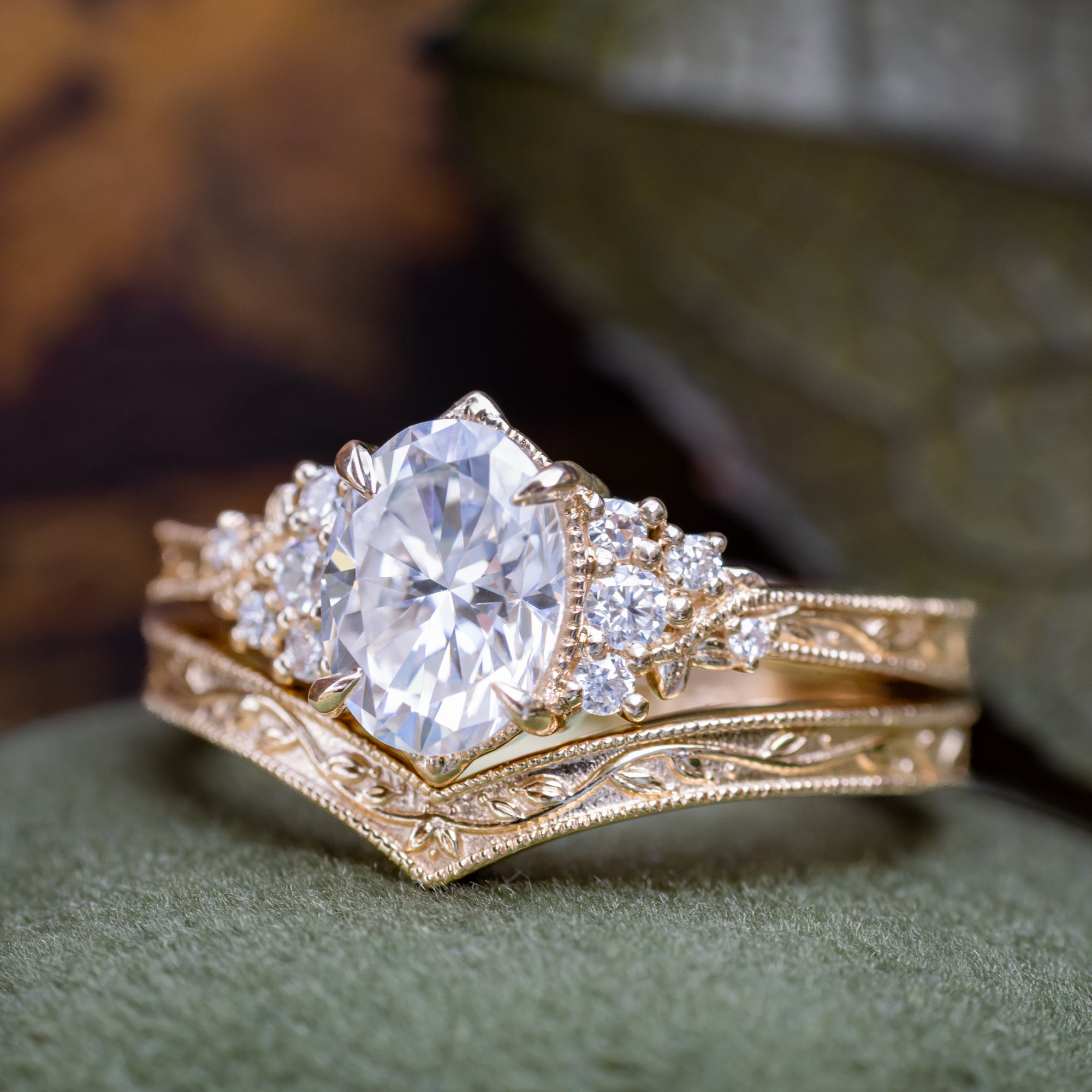A gold engagement ring with an oval Moissanite and diamond accents, shown resting on a textured green surface with blurred background.