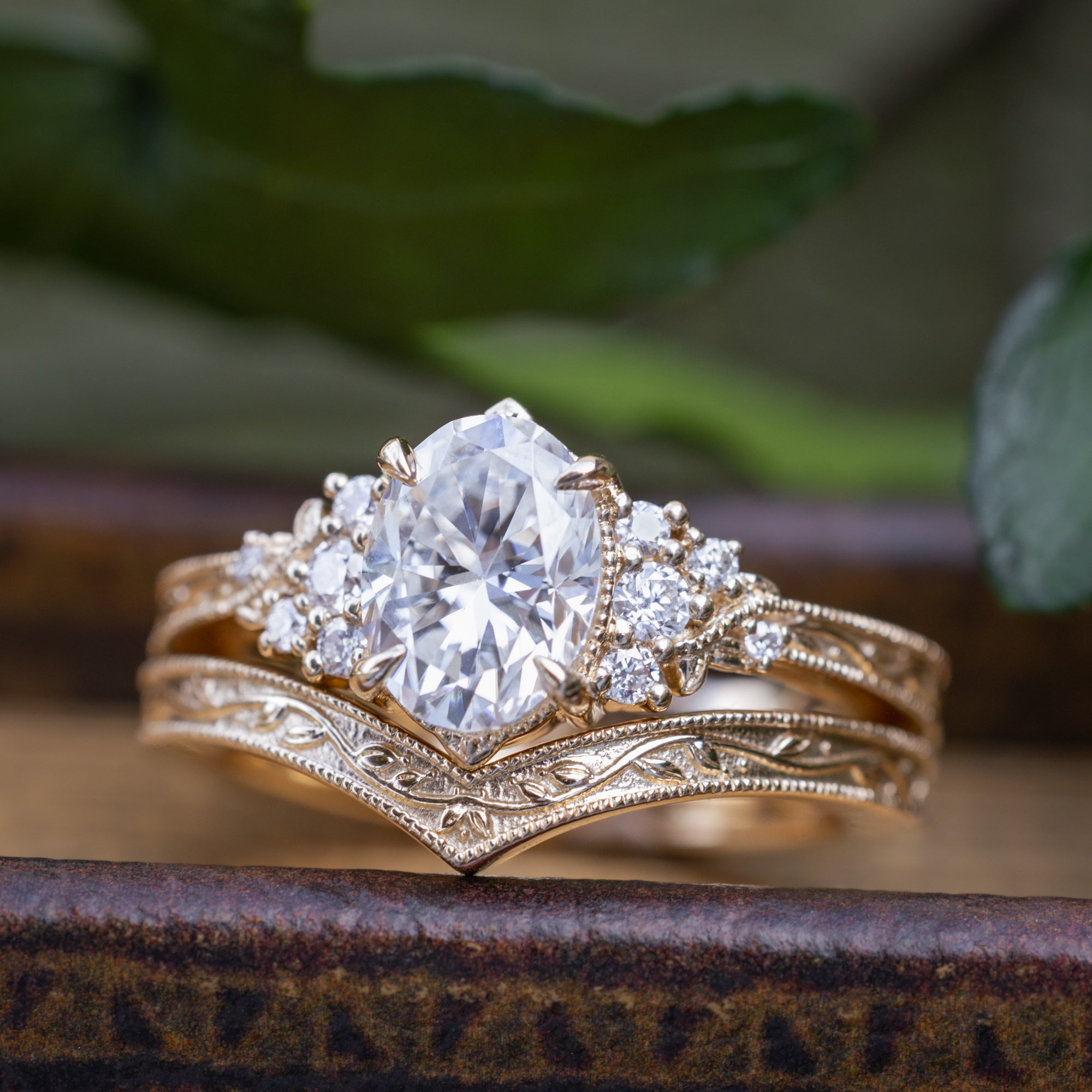 A gold engagement ring with an oval Moissanite and diamond accents, shown resting on a rustic wooden surface with blurred green leaves in the background.