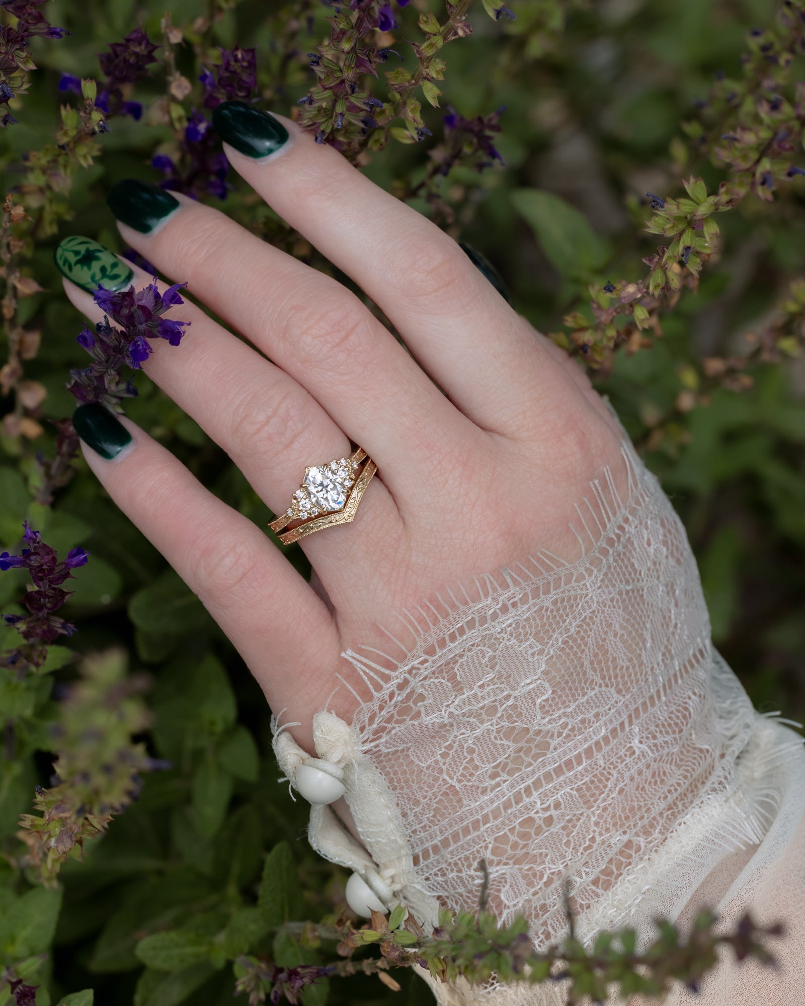 A close-up of a hand wearing a gold ring with a moissanite, styled with a lace sleeve, against a background of green foliage and purple flowers.