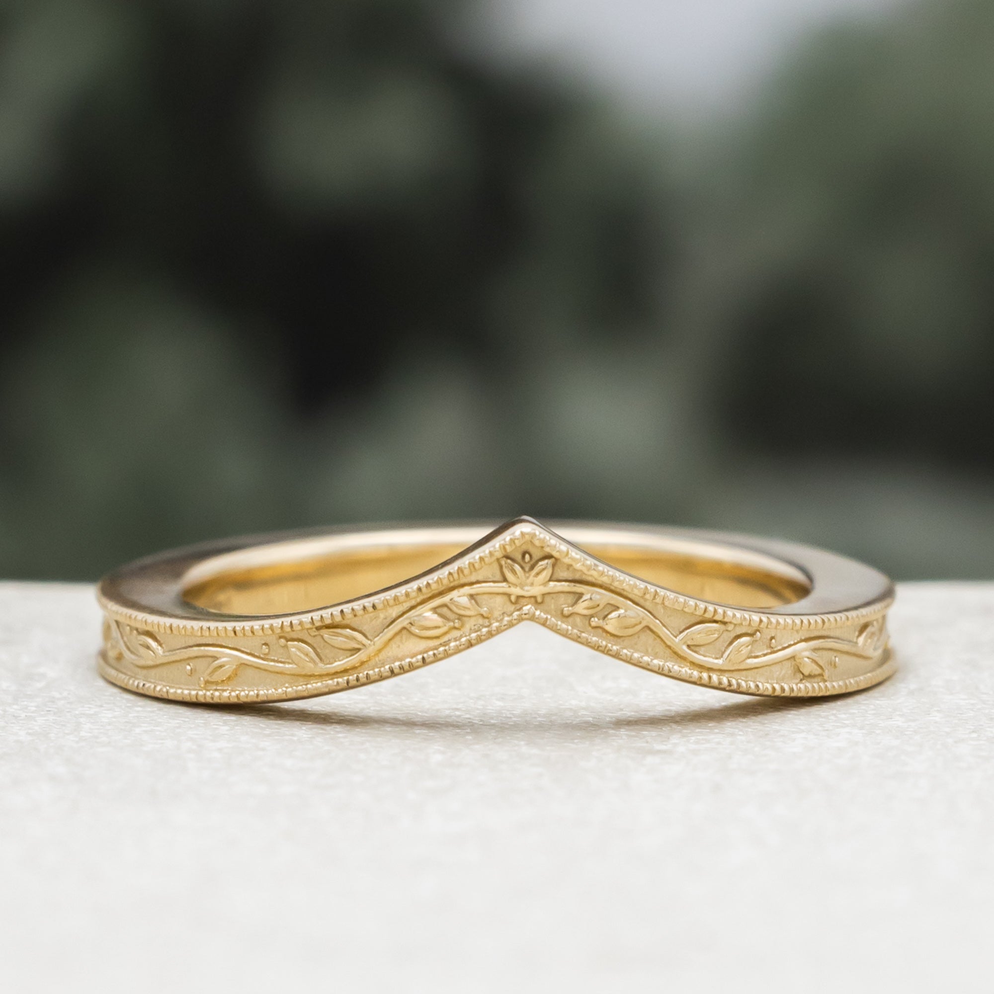 A gold V-shaped ring with leaf filigree, on a textured surface against a blurred green background