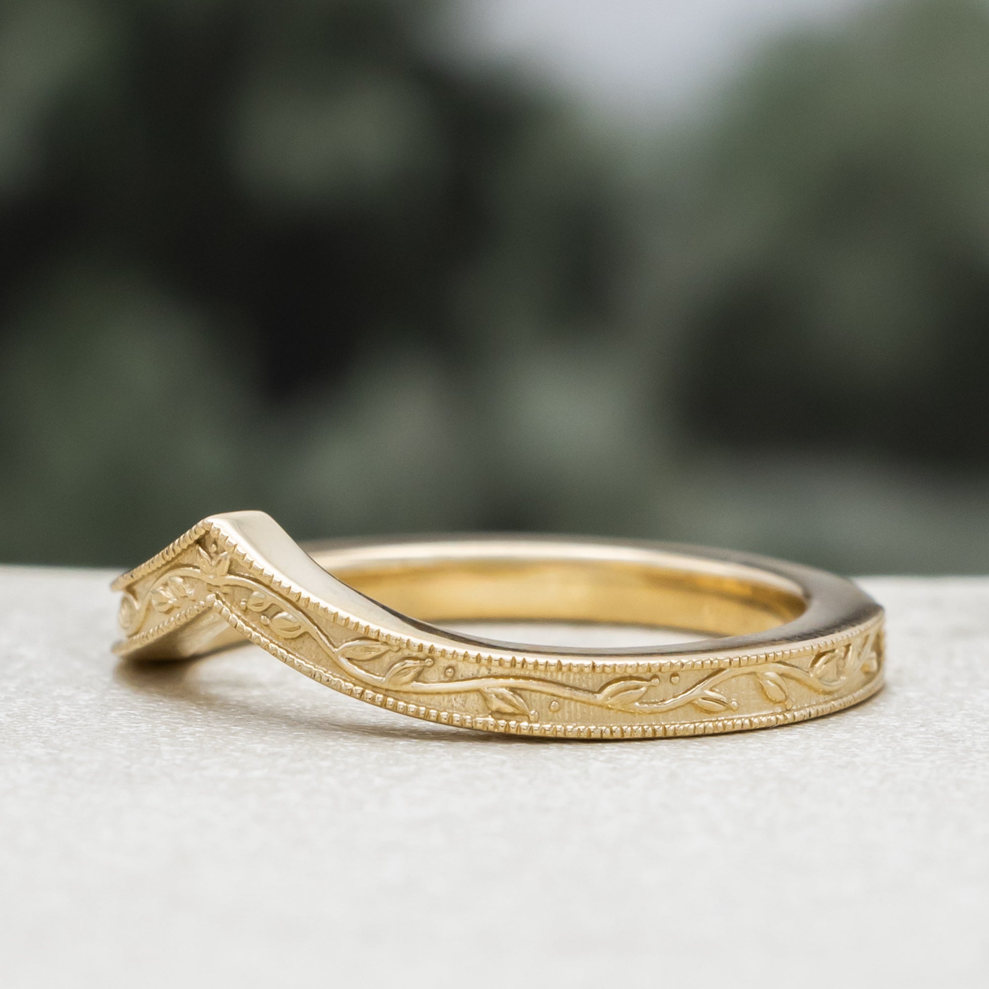 A gold V-shaped ring with leaf filigree, on a textured surface against a blurred green background