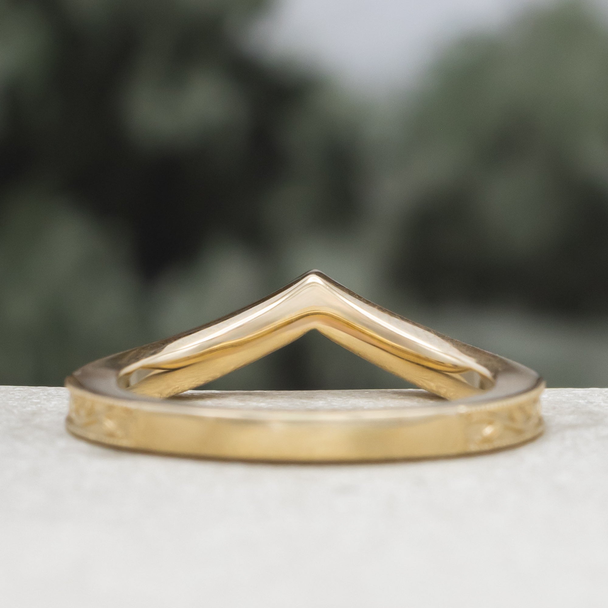 A gold V-shaped ring with leaf filigree, on a textured surface against a blurred green background