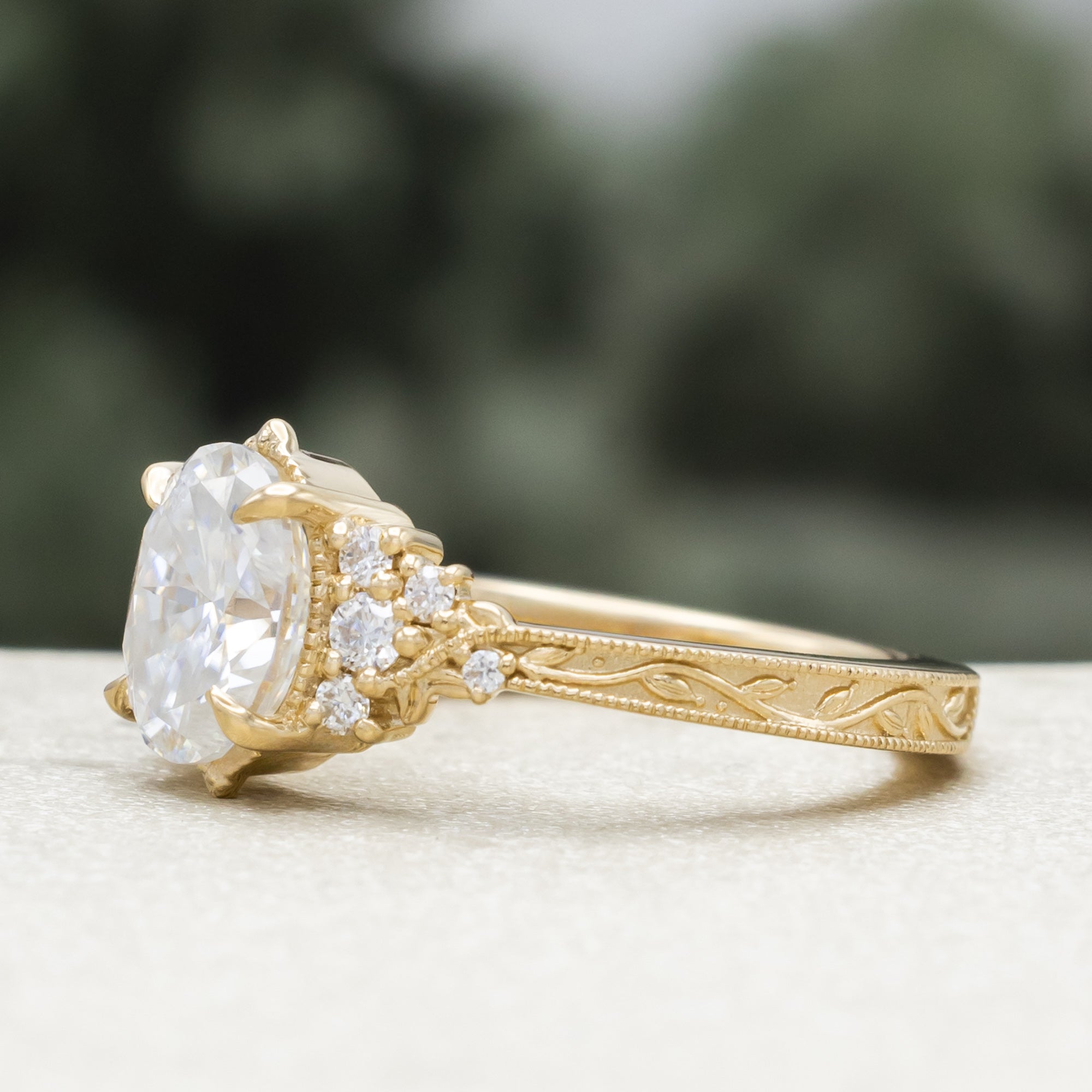 A gold engagement ring with an oval Moissanite and diamond accents, against a blurred green background