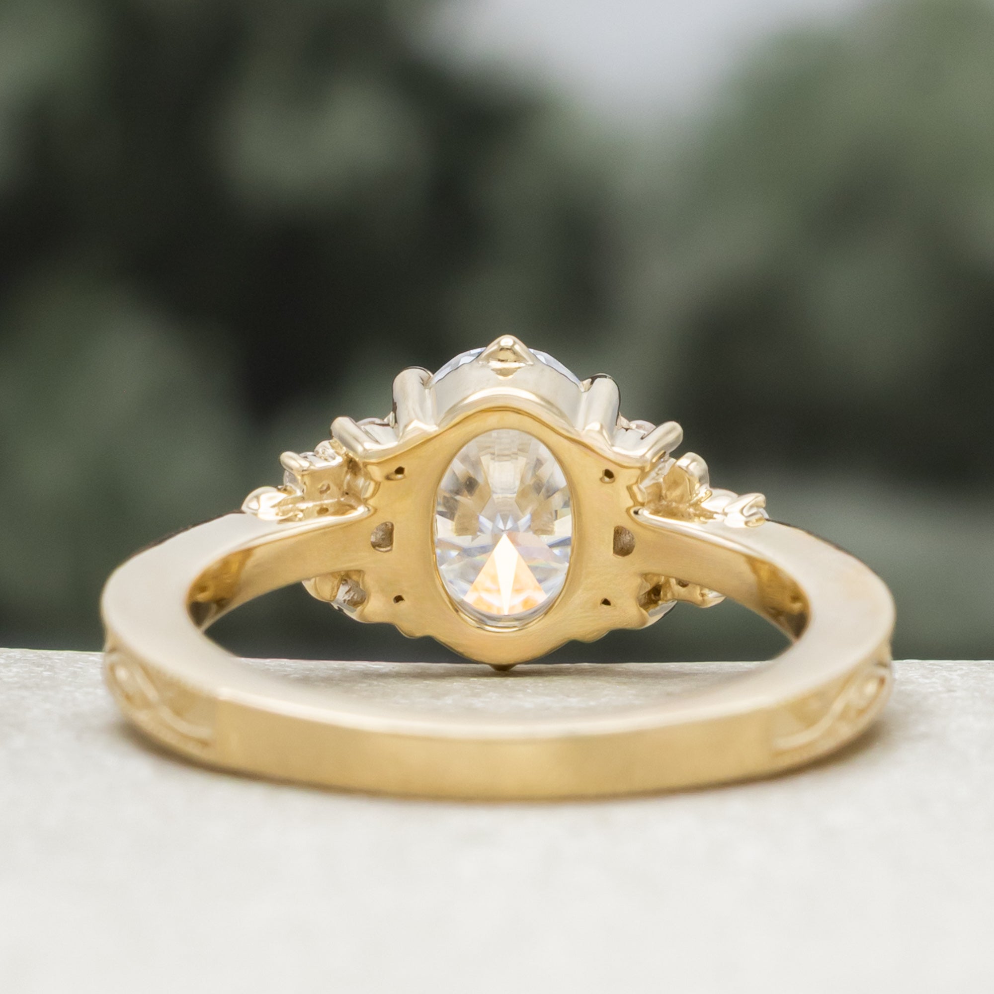 A gold engagement ring with an oval Moissanite and diamond accents, against a blurred green background