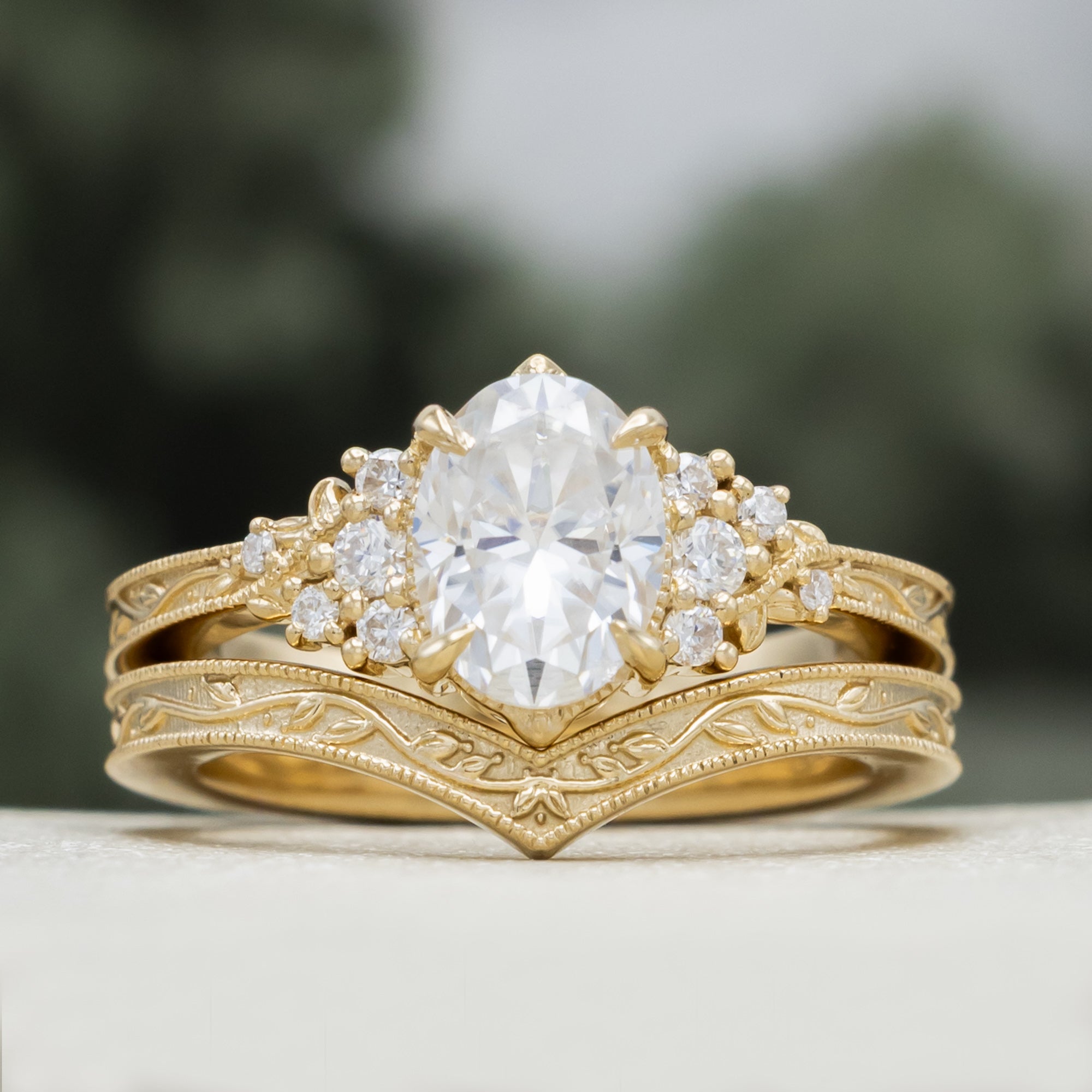 A gold engagement ring and tracer with an oval Moissanite and diamond accents, against a blurred green background