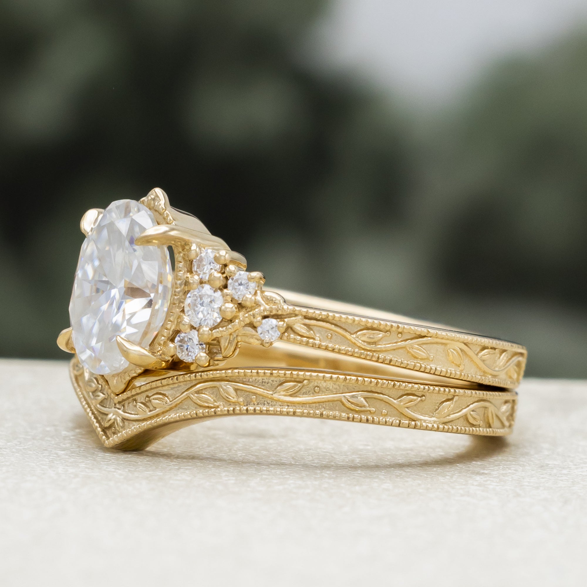 A gold engagement ring and tracer with an oval Moissanite and diamond accents, against a blurred green background