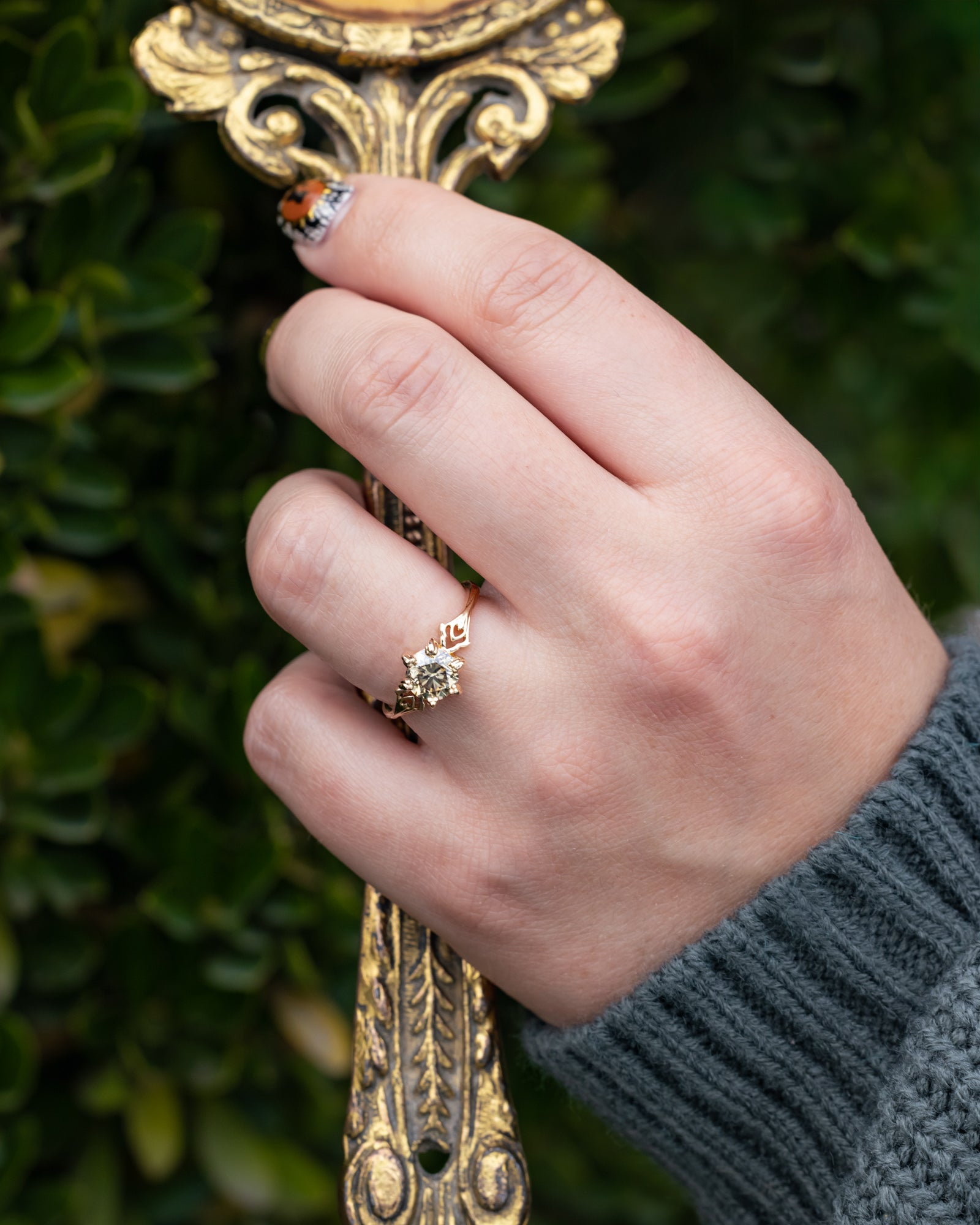 Hand wearing a ring with a decorative gold object against a green leafy background