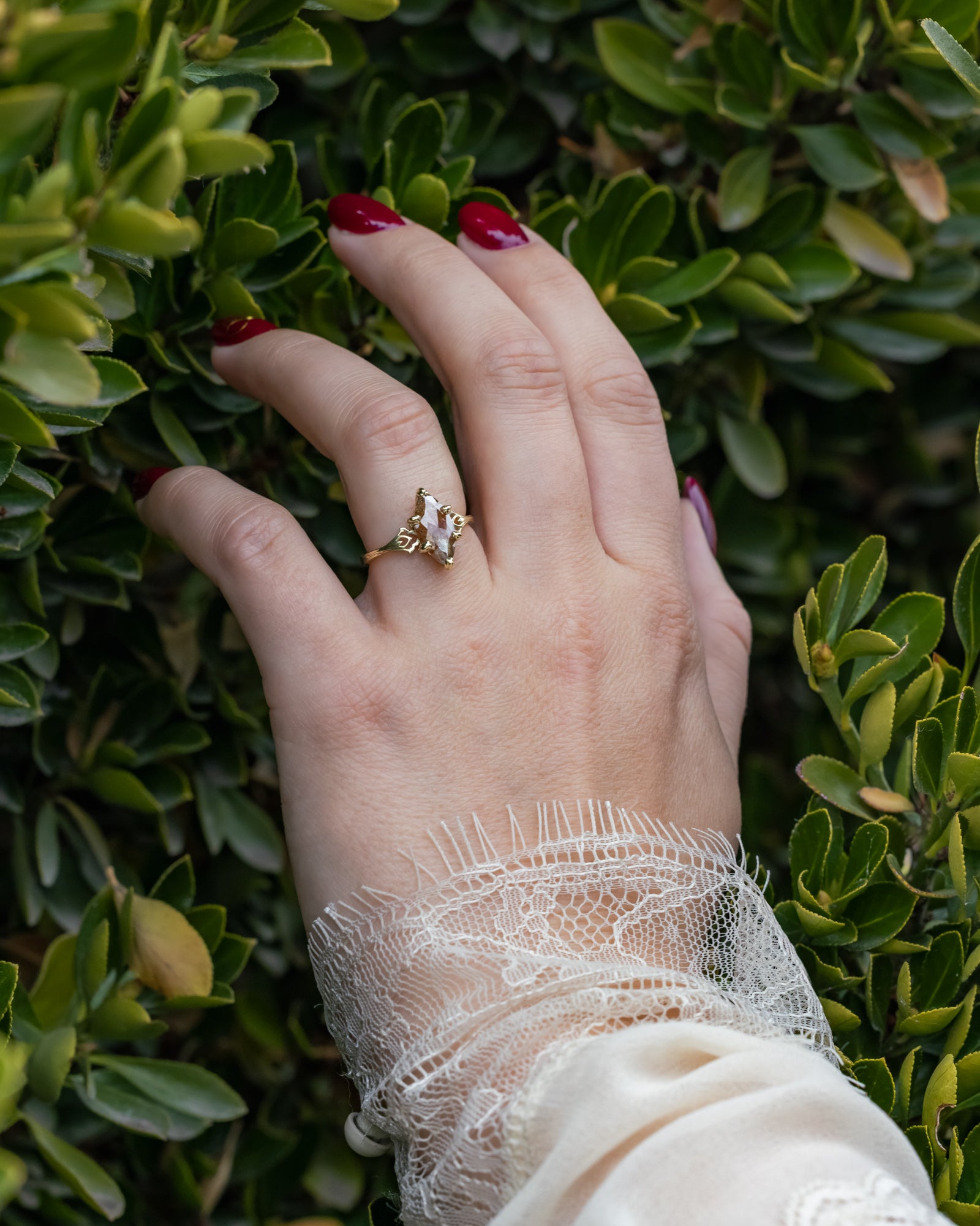 Hand wearing a gold ring with a salt & pepper diamond, against a green leafy background