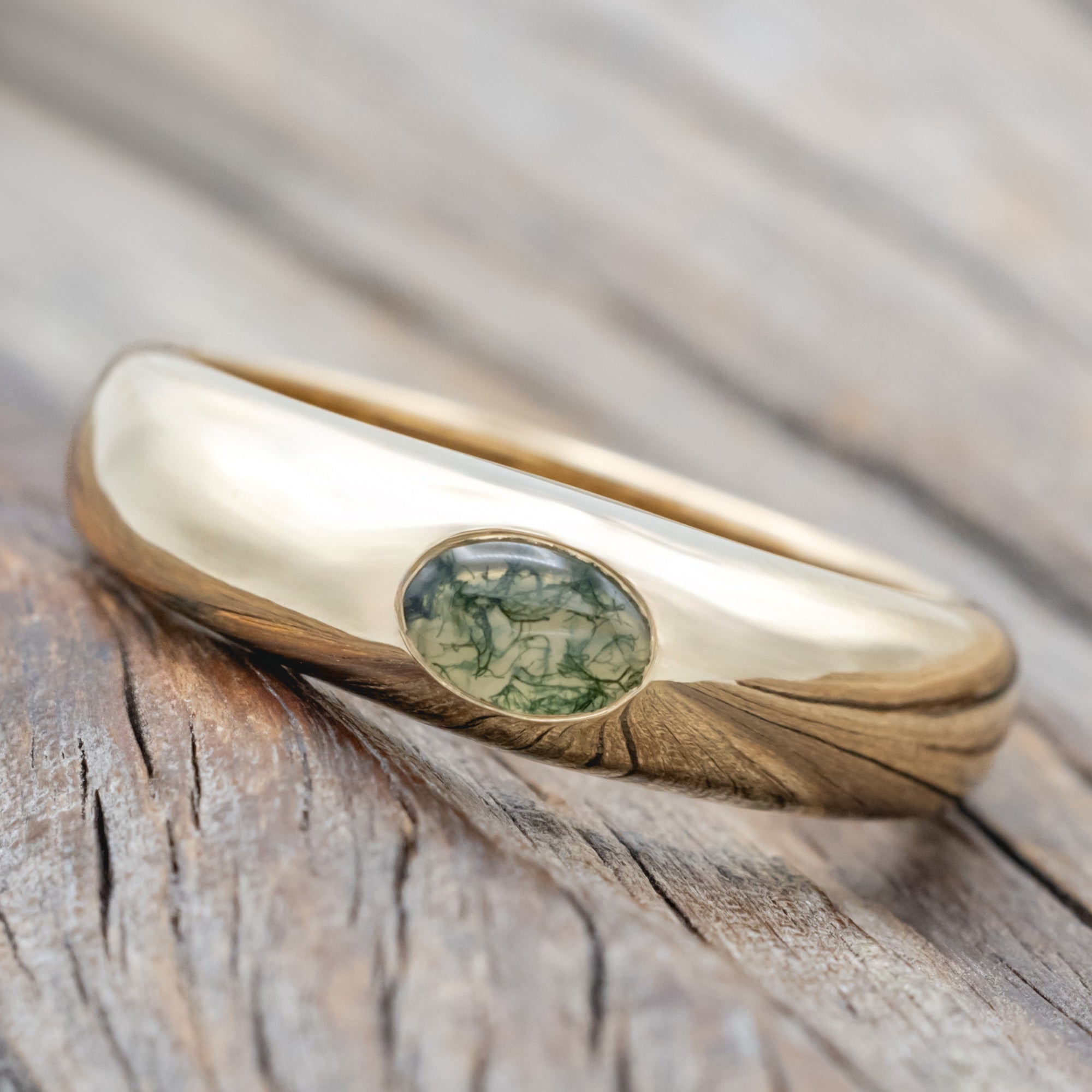 A gold ring with a bezel set moss agate displayed on a wooden surface.