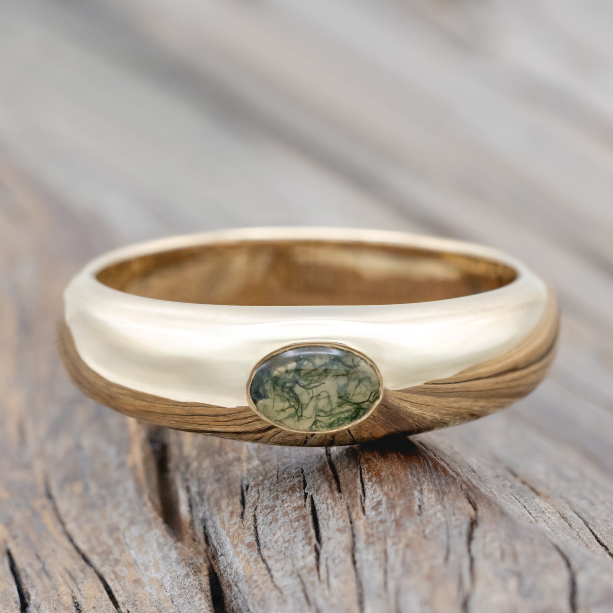 A gold ring with a bezel set moss agate displayed on a wooden surface.