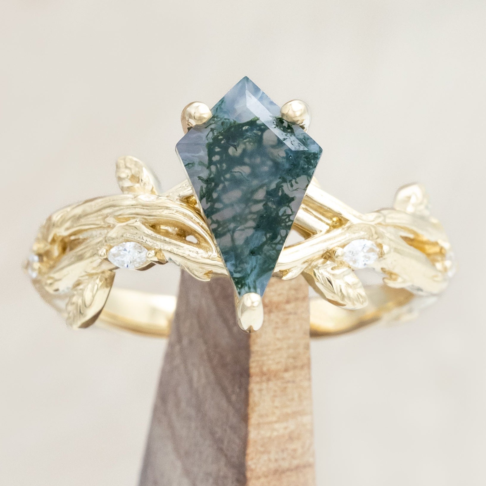 Gold ring with a moss agate with vine-style band and diamond accents on a wooden stand against a light background