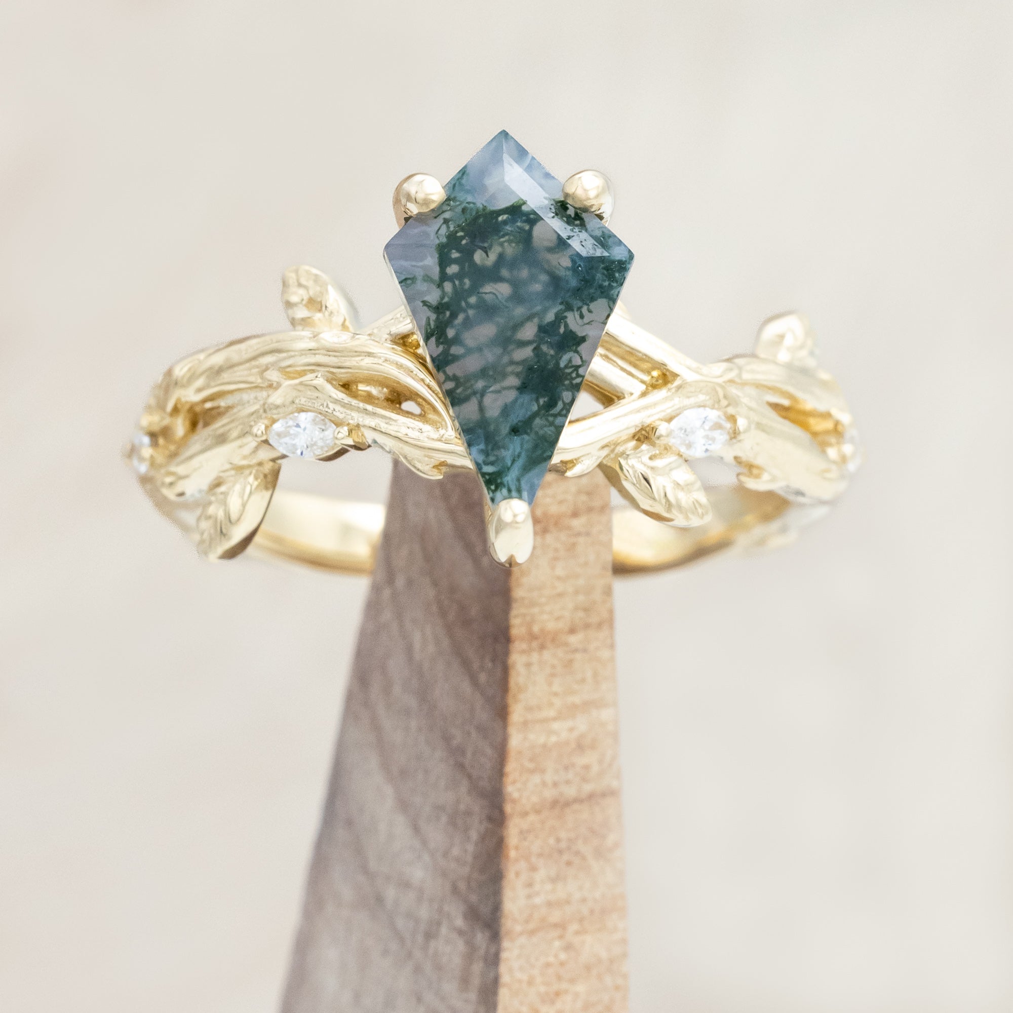 Gold ring with a moss agate with vine-style band and diamond accents on a wooden stand against a light background