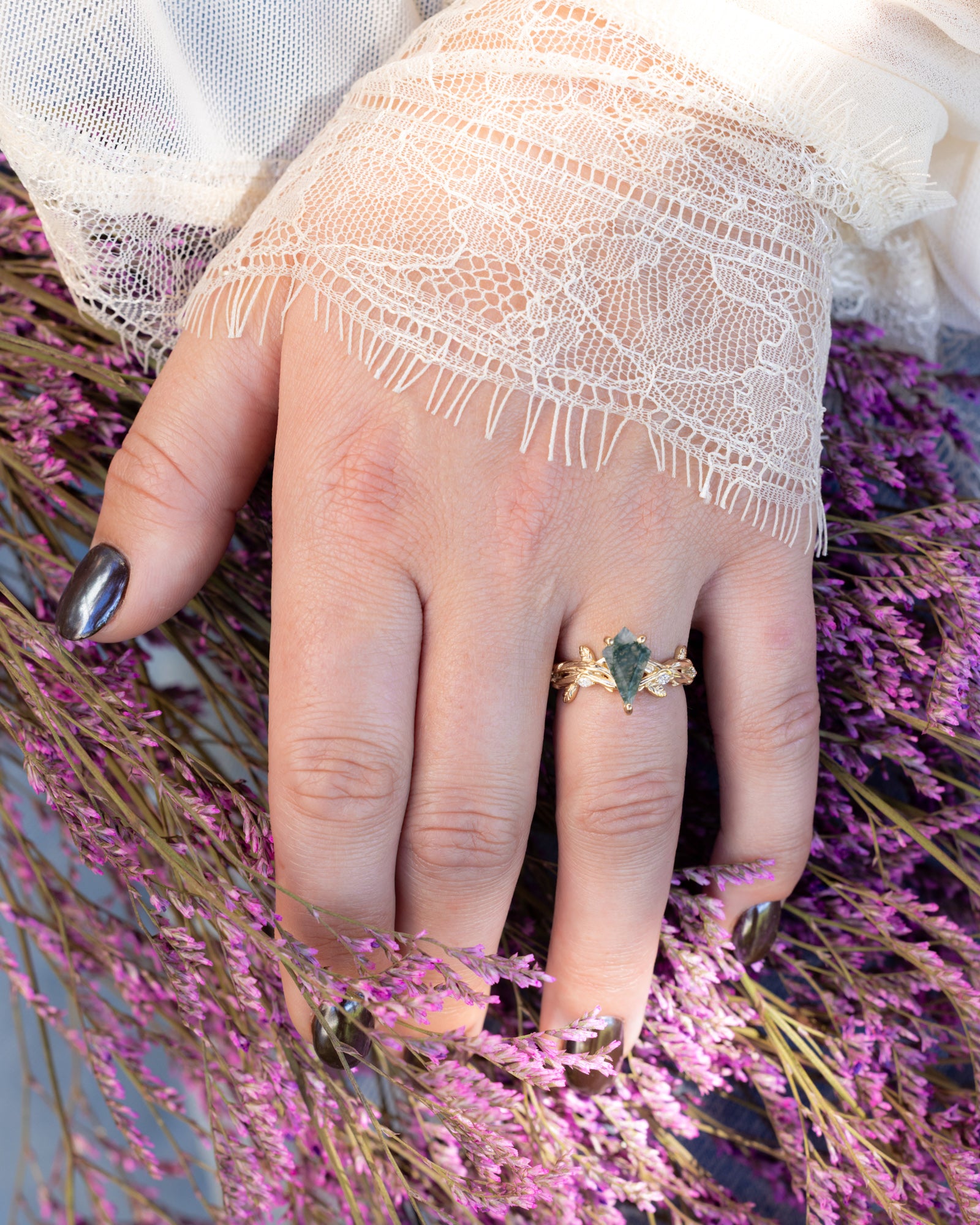 Hand wearing a gold ring with moss agate on a background of purple flowers