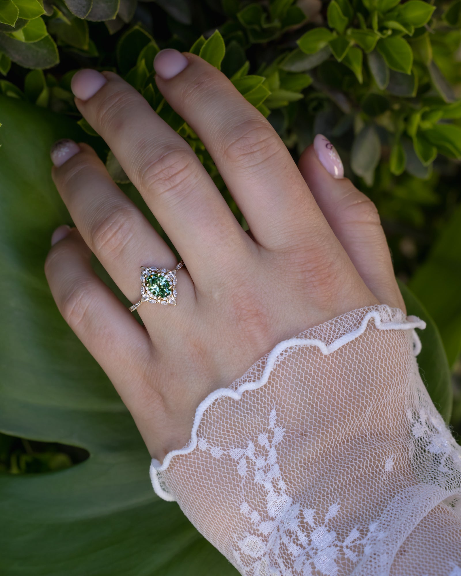 Hand wearing a diamond ring with green lab diamond against a green leafy background