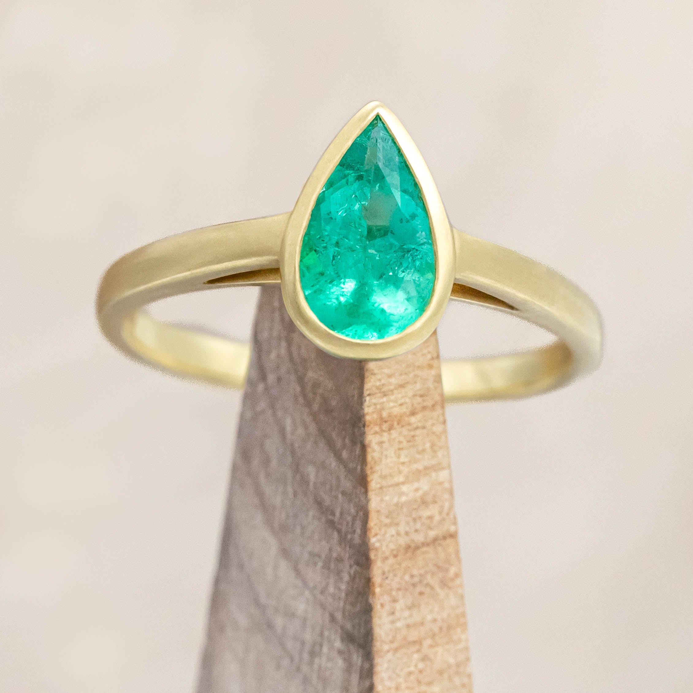 Gold ring with a pear cut emerald on a wooden stand against a beige background