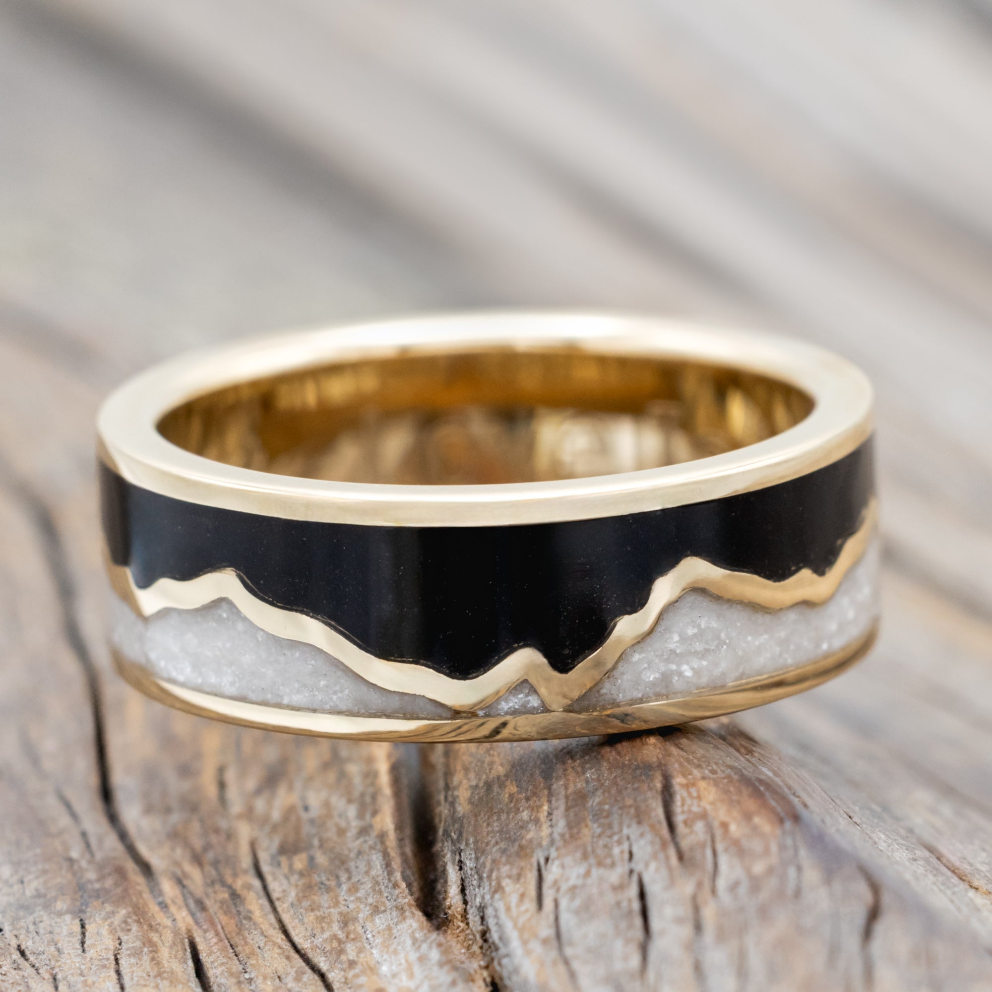 A gold ring with jet stone sky and diamond dust inlays with gold mountains between the two on a wooden surface