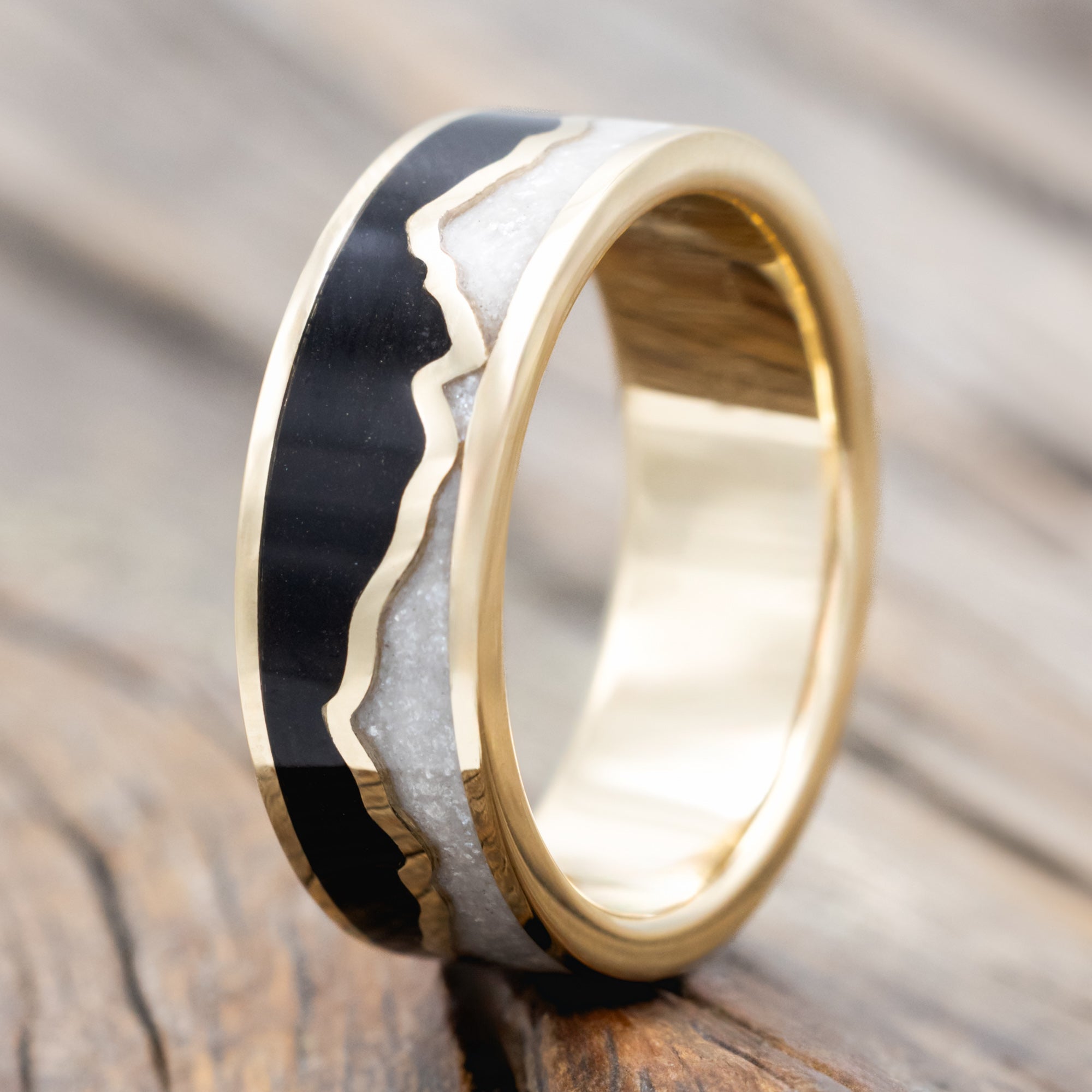 A gold ring with jet stone sky and diamond dust inlays with gold mountains between the two on a wooden surface