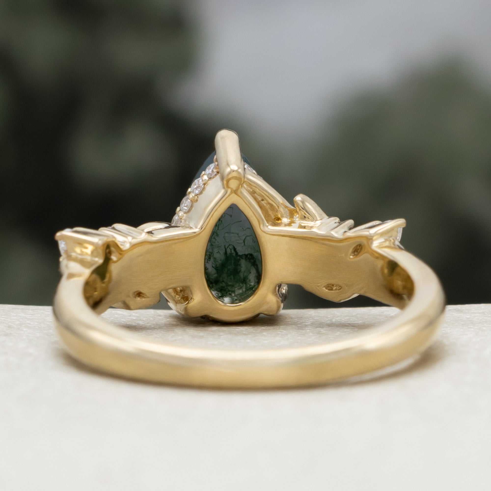 A gold ring featuring a pear cut moss agate flanked by delicate gold leaf-like branches with diamond accents, set against a blurred dark green background.