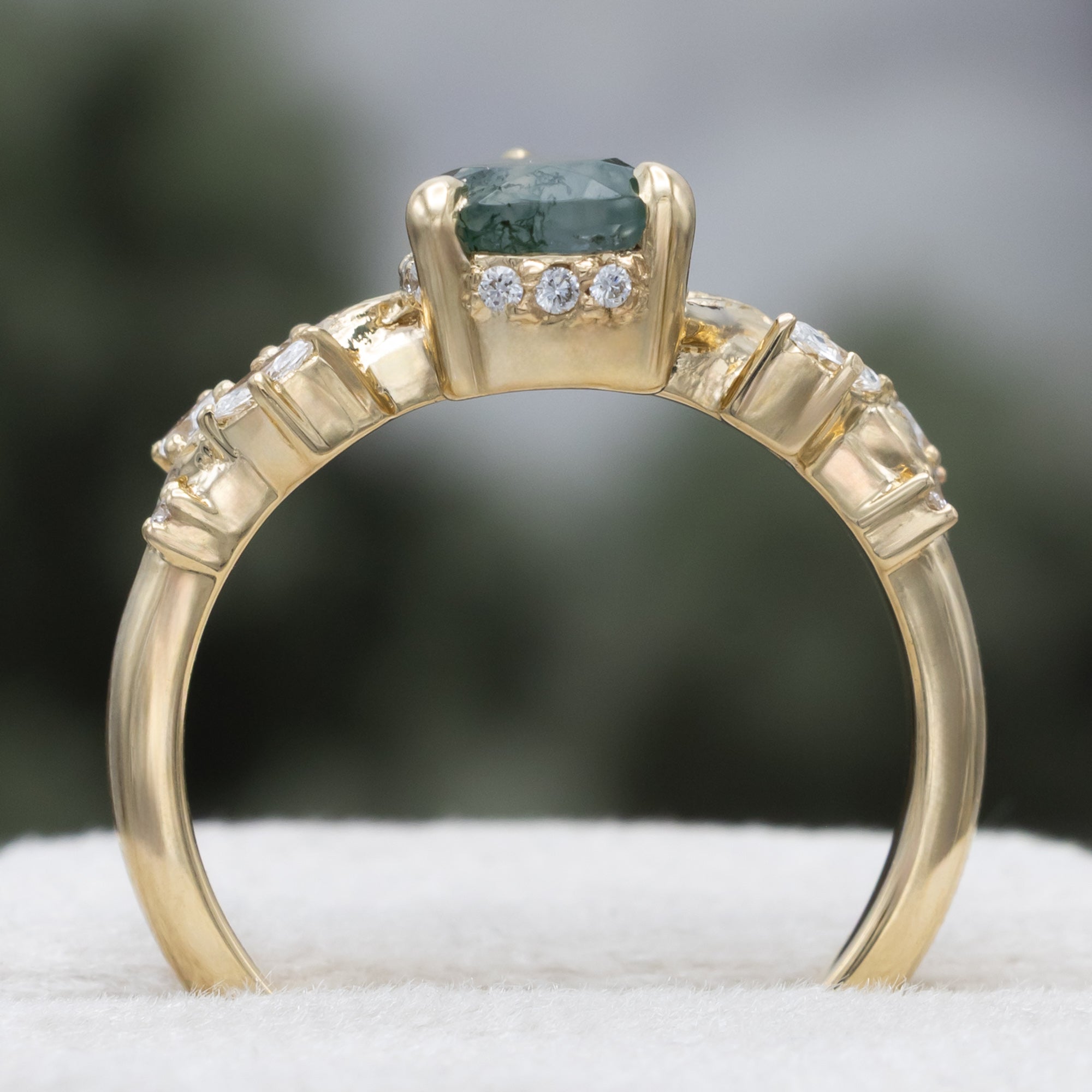 A gold ring featuring a pear cut moss agate flanked by delicate gold leaf-like branches with diamond accents, set against a blurred dark green background.