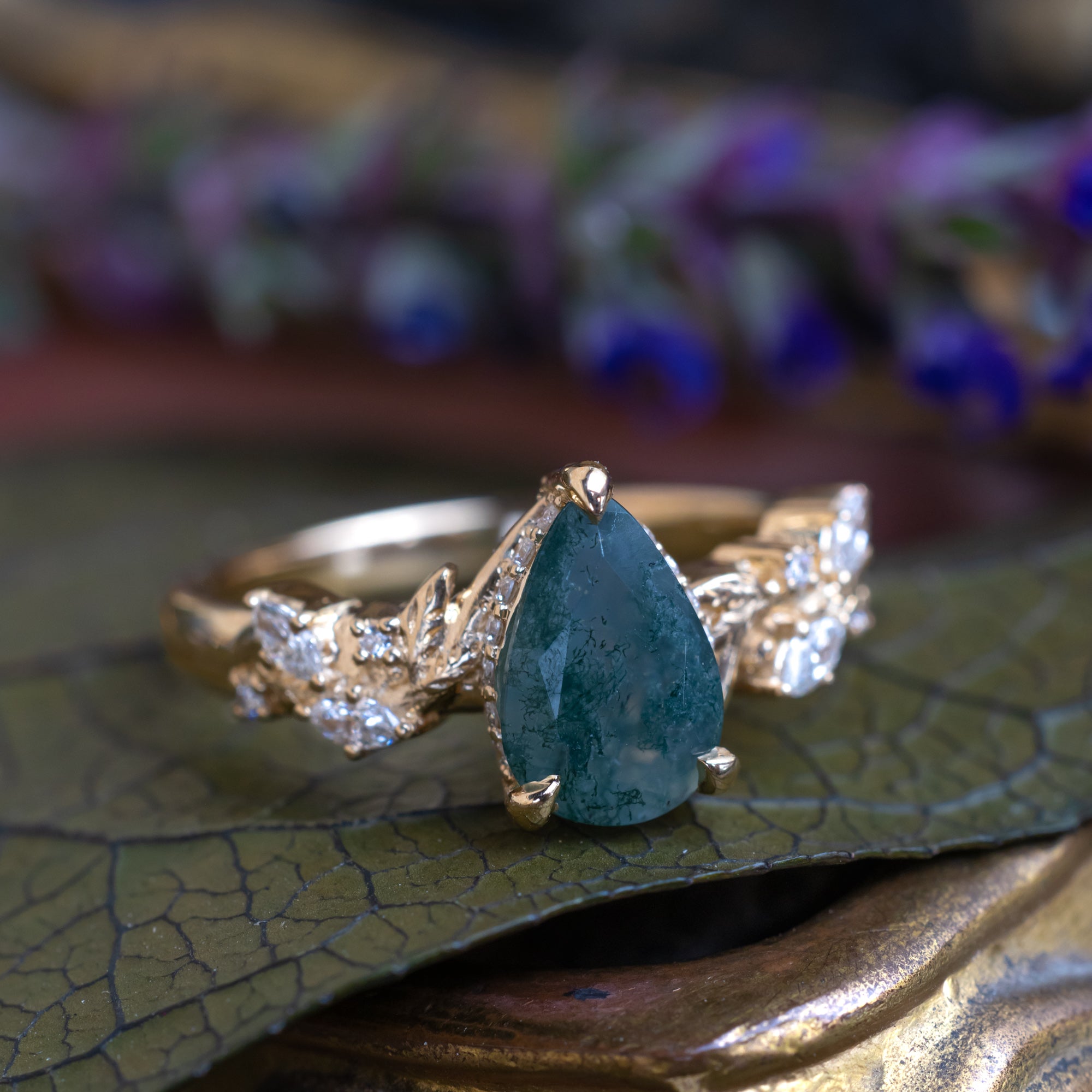 A gold ring featuring a pear-cut moss agate flanked by delicate gold leaf-like branches with diamond accents resting on a textured green leaf with a blurred purple floral background.