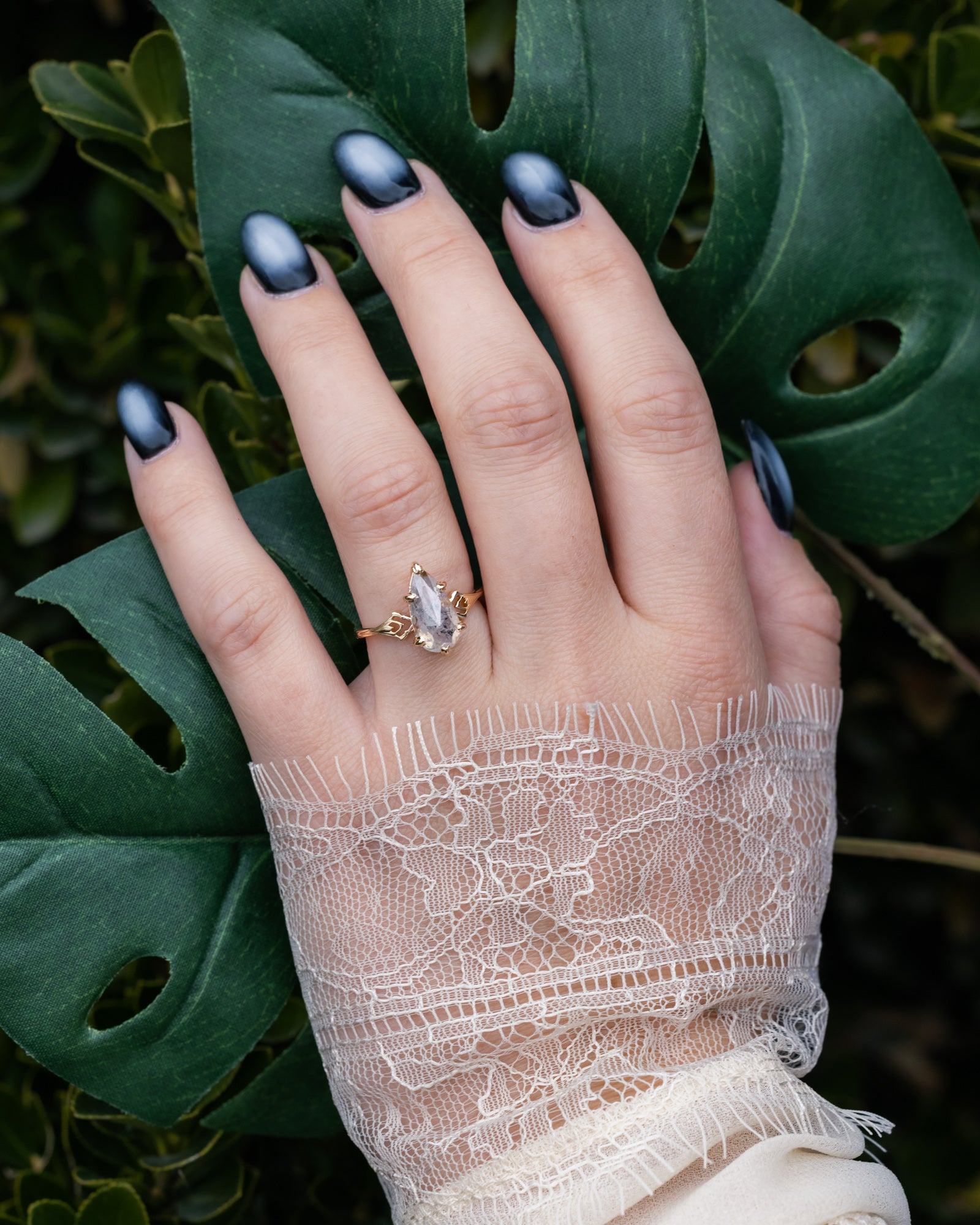 Hand with dark nail polish and salt & pepper diamond ring, wearing a lace sleeve against a leafy background
