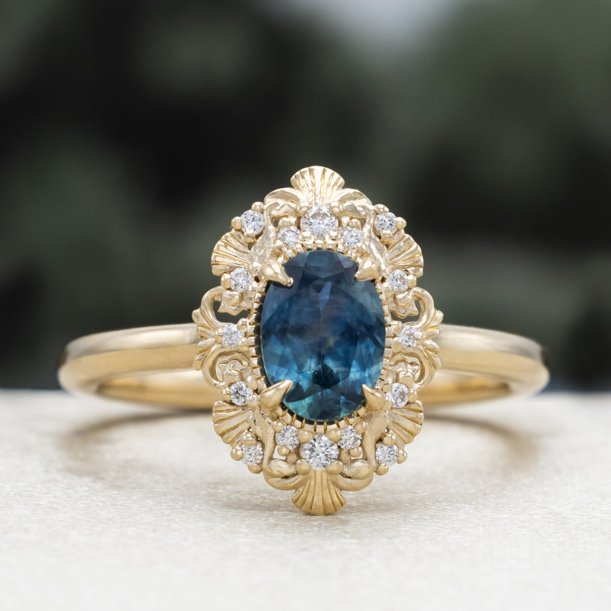 A gold ring with Montana sapphire center stone and diamond accents against a blurred green background