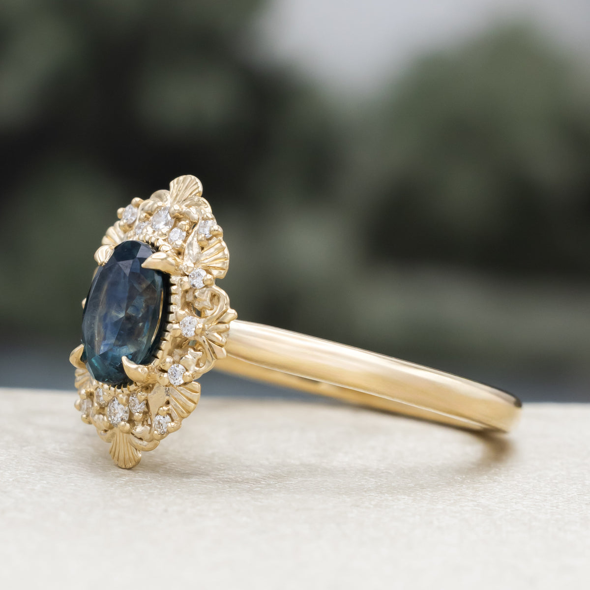 A gold ring with Montana sapphire center stone and diamond accents against a blurred green background